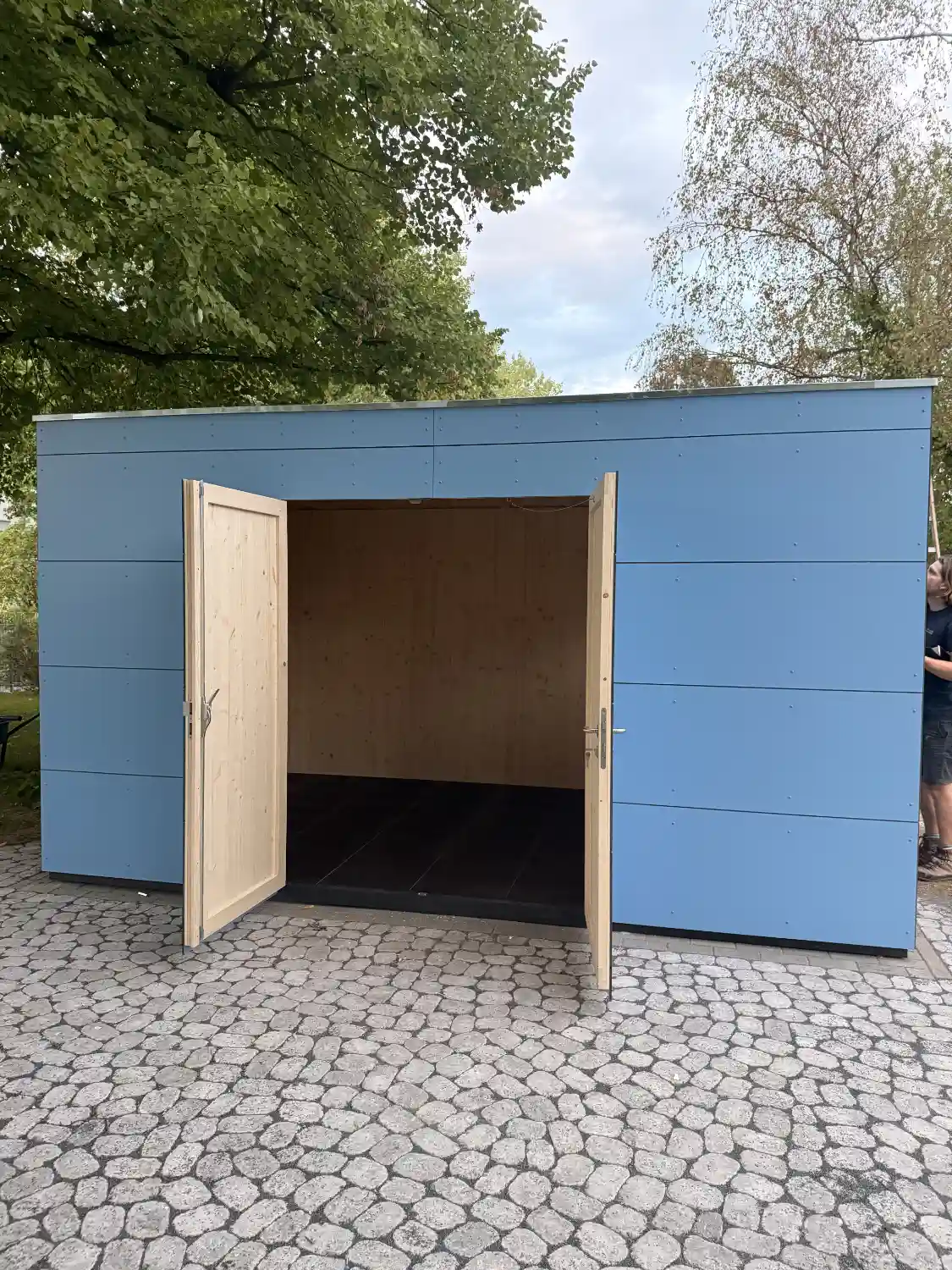 Blick auf Blaues Gerätehaus mit Flachdach, offen stehender Doppelflügeltür und blauer HPL-Fassade, im Hintergrund grüne Bäume