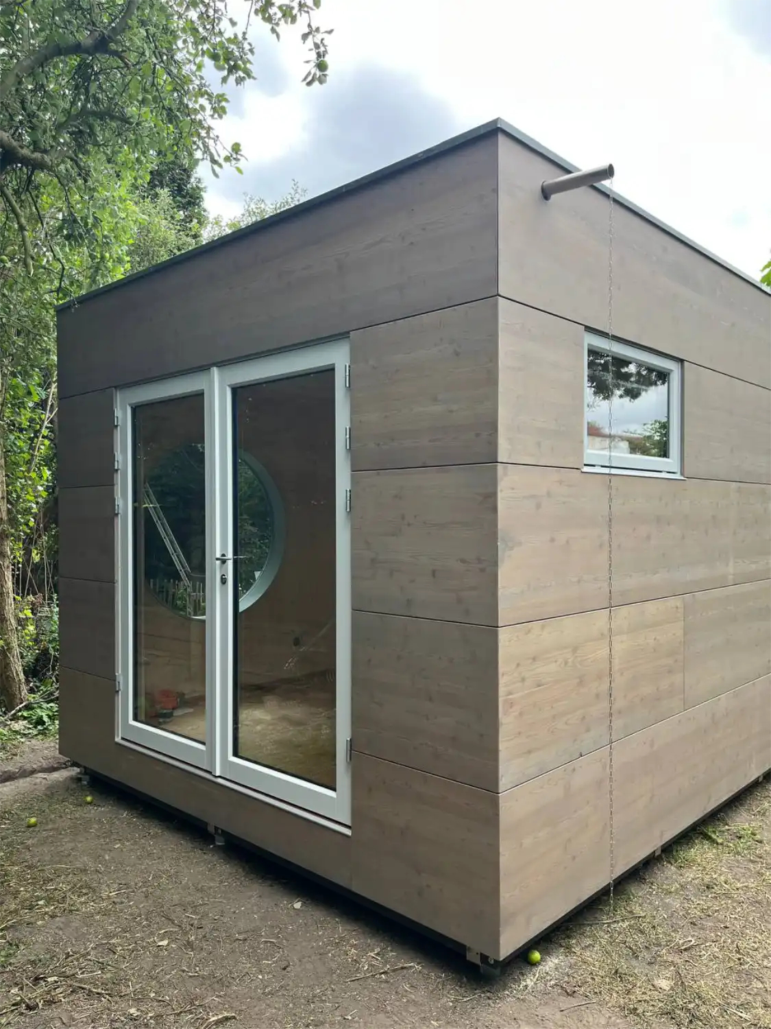 Blick auf Seite eines großen Gartenbüros mit Flachdach, großer Glastür, Fenster und grau lasierter Lärche Fassade