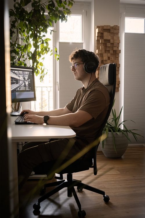 Mann mit Kopfhörern und Brille arbeitet an einem Computer in einem hellen Raum mit Pflanzen und schallabsorbierenden Holzwürfeln an der Wand.