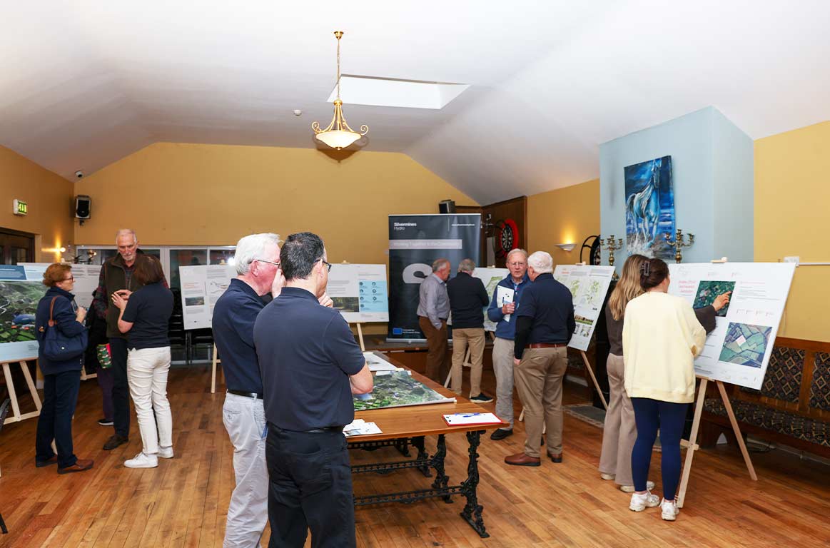 People in a room viewing and discussing maps and informational boards displayed on easels during a public consultation event.