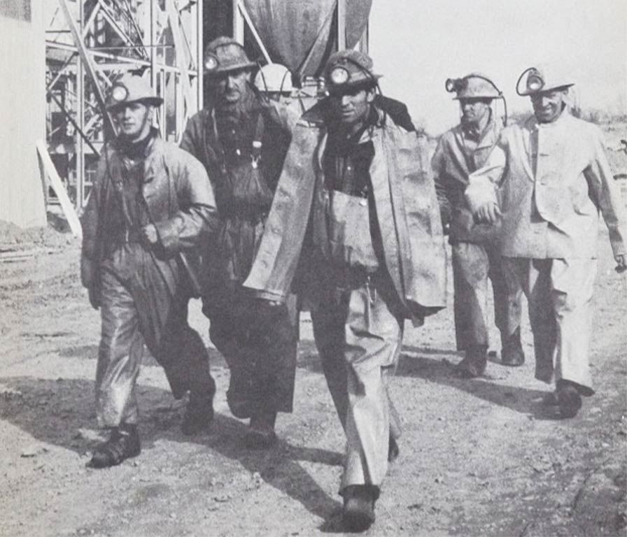 Black and white photo of five miners walking outdoors wearing helmets with lamps and protective gear.