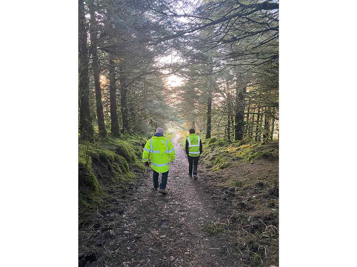 Two people in high-visibility jackets walking on a forest path surrounded by trees and moss.