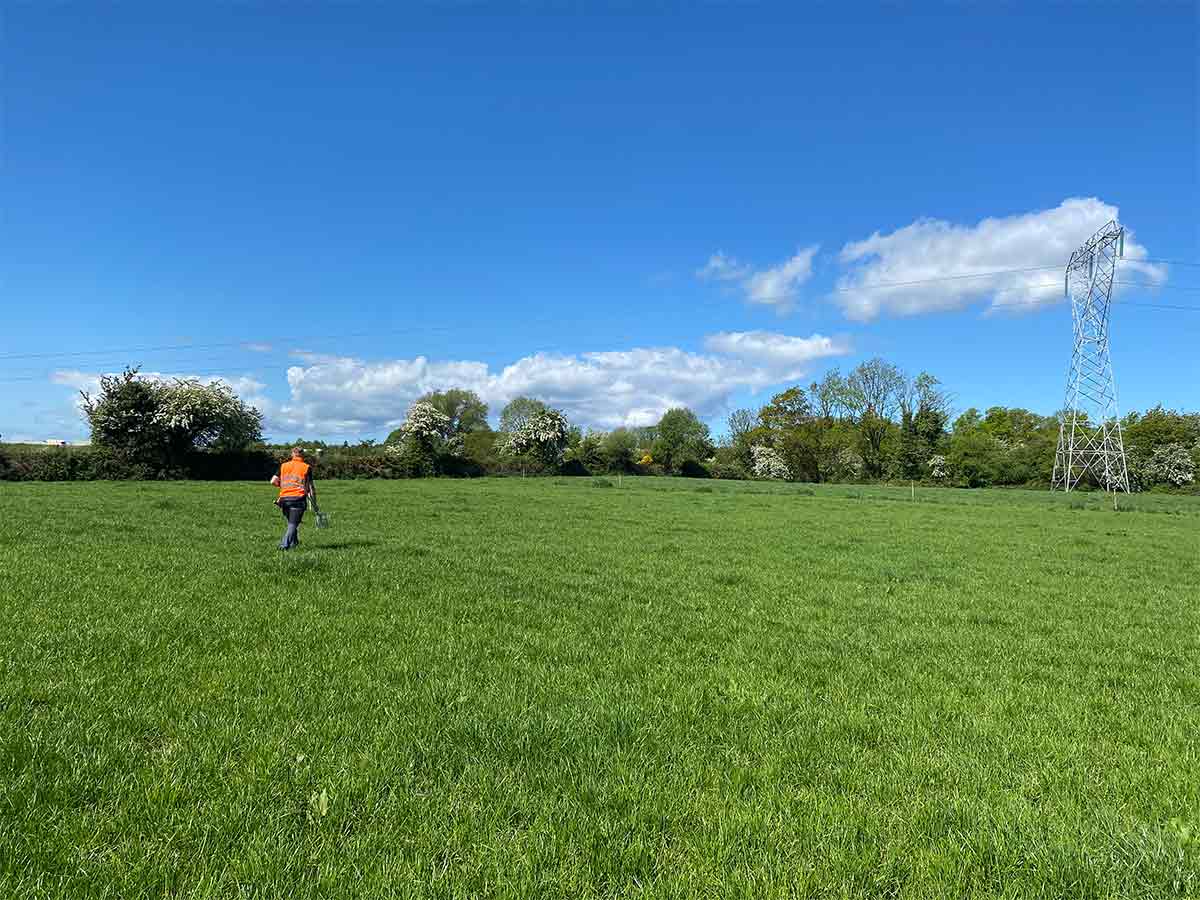 Person in orange vest walking through green grassy field under blue sky with power line tower.