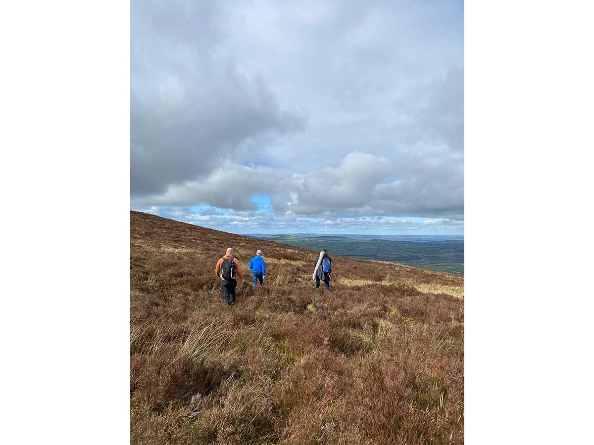 Three hikers walking through a grassy hill under a cloudy blue sky.