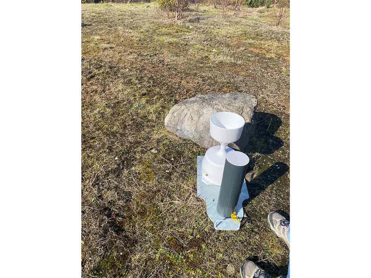 White weather station instrument on ground near a large rock and a person’s sneaker.