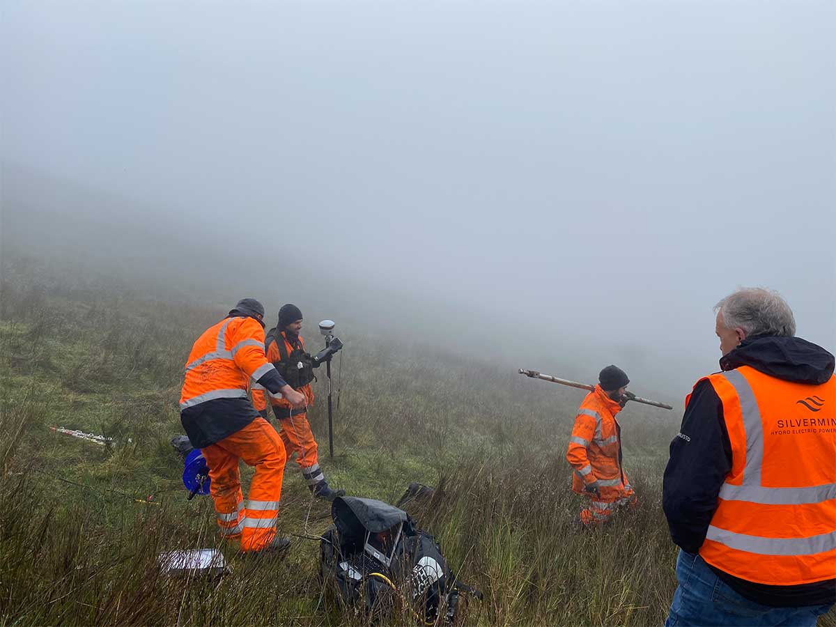 Four workers in orange reflective gear conducting surveying in foggy grassy field.