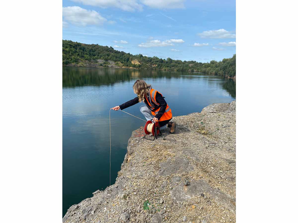 Person in orange safety vest kneeling by water, lowering a measuring line into the lake.