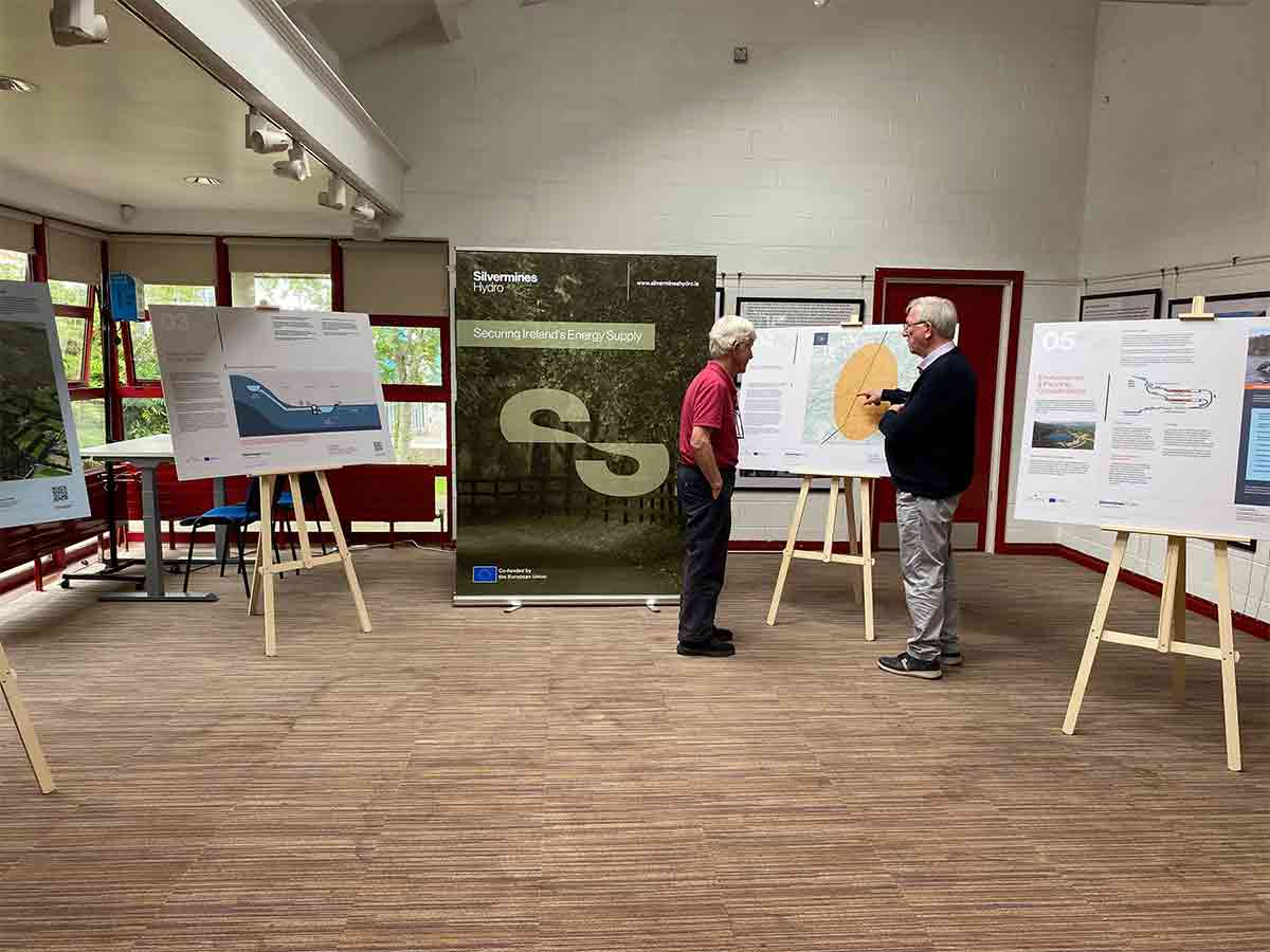 Two men discuss maps and charts in a well-lit room with informational boards on easels.