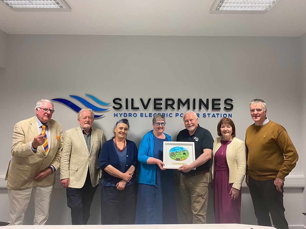 Seven people stand smiling in a room under a Silvermines Hydro Electric Power Station sign holding a framed artwork.