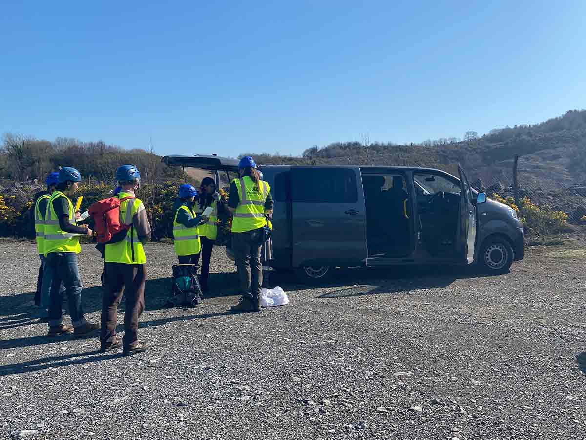 Group in yellow safety vests and blue helmets standing near a van in a gravel area on a sunny day.