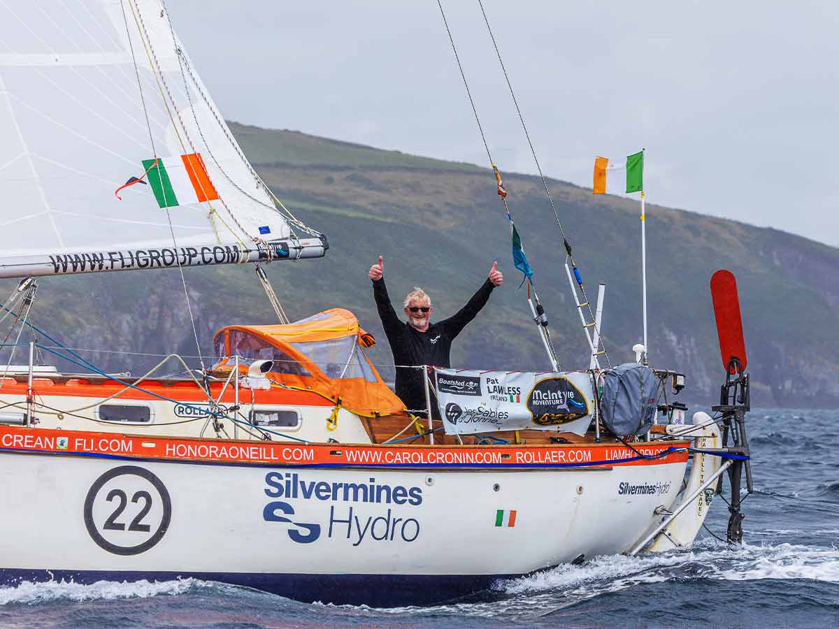 Man on small sailing boat with Irish flags, giving two thumbs up on open water near cliffs.