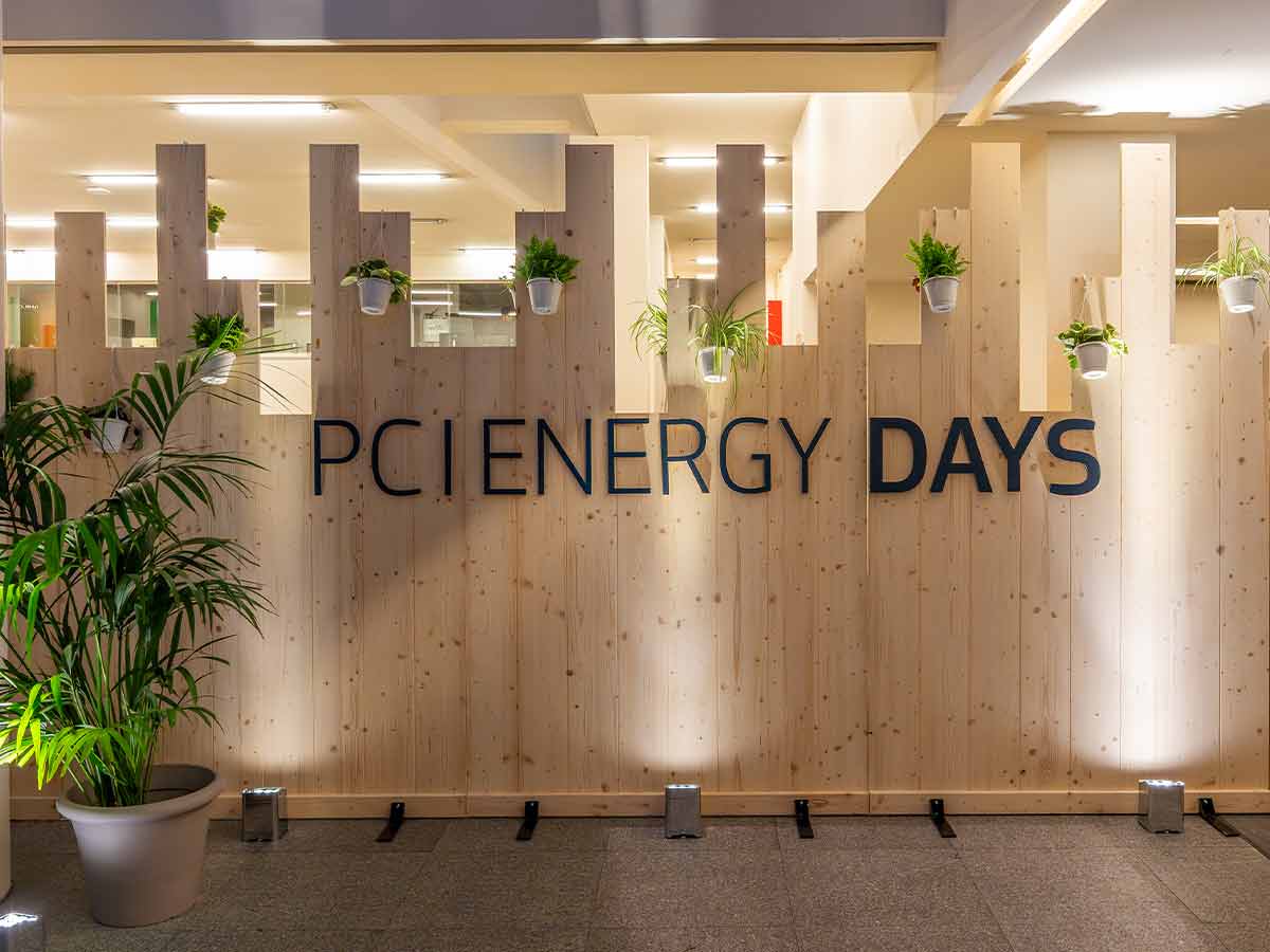 Wooden wall with 'PCI ENERGY DAYS' text and potted plants hanging and placed in front.