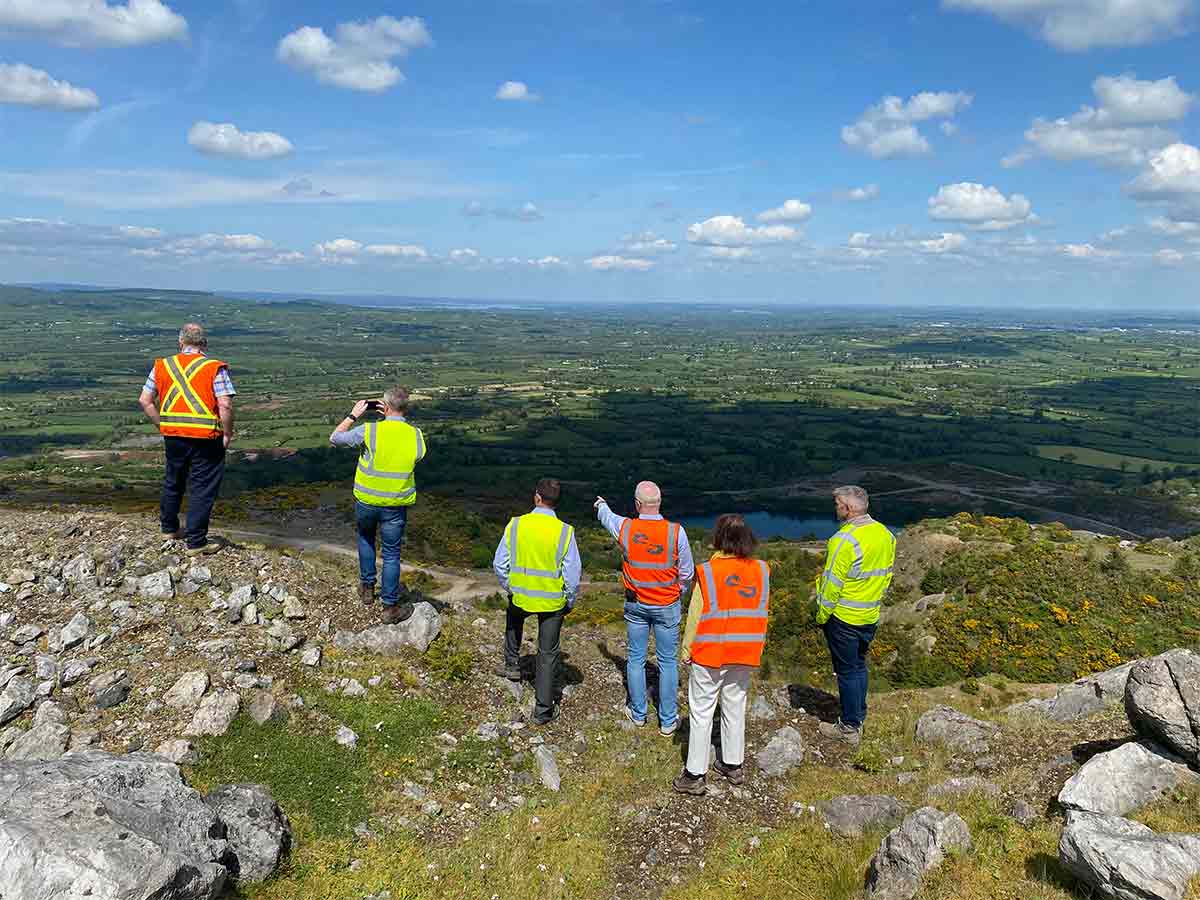 Six people in safety vests stand on rocky hilltop overlooking expansive green valley under blue sky.