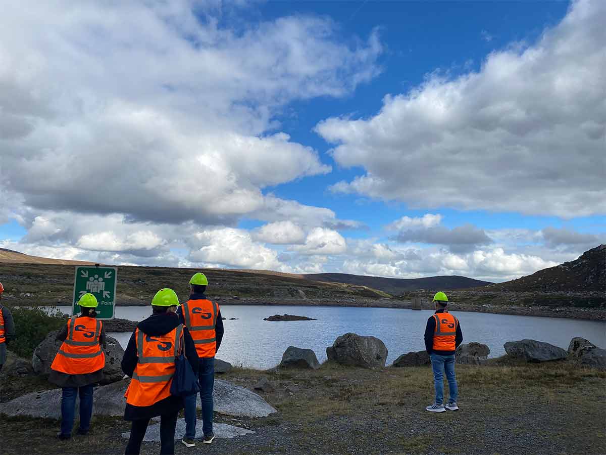 Four people in orange safety vests and green helmets stand by a lake with rocky hills under a cloudy sky.