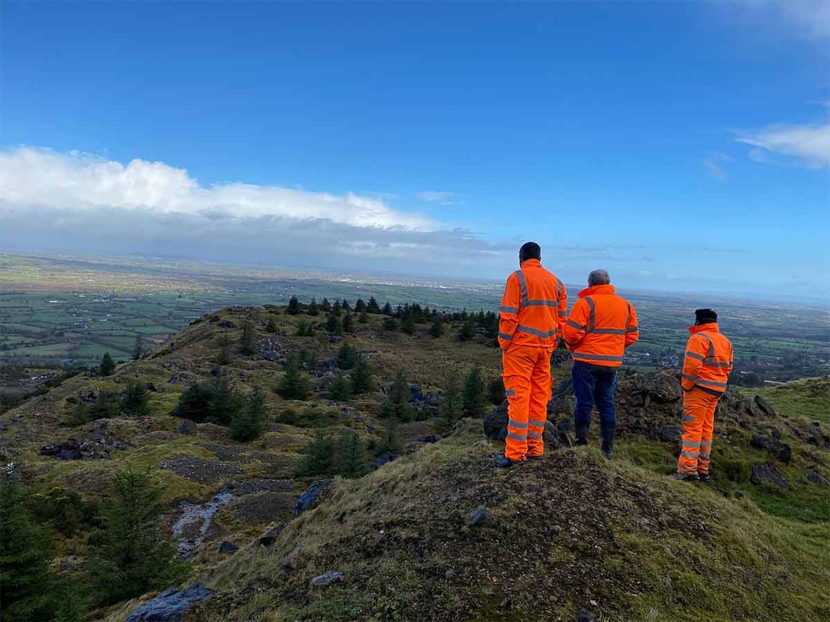 Three people in orange safety gear stand on a hilltop overlooking green fields and a blue sky.