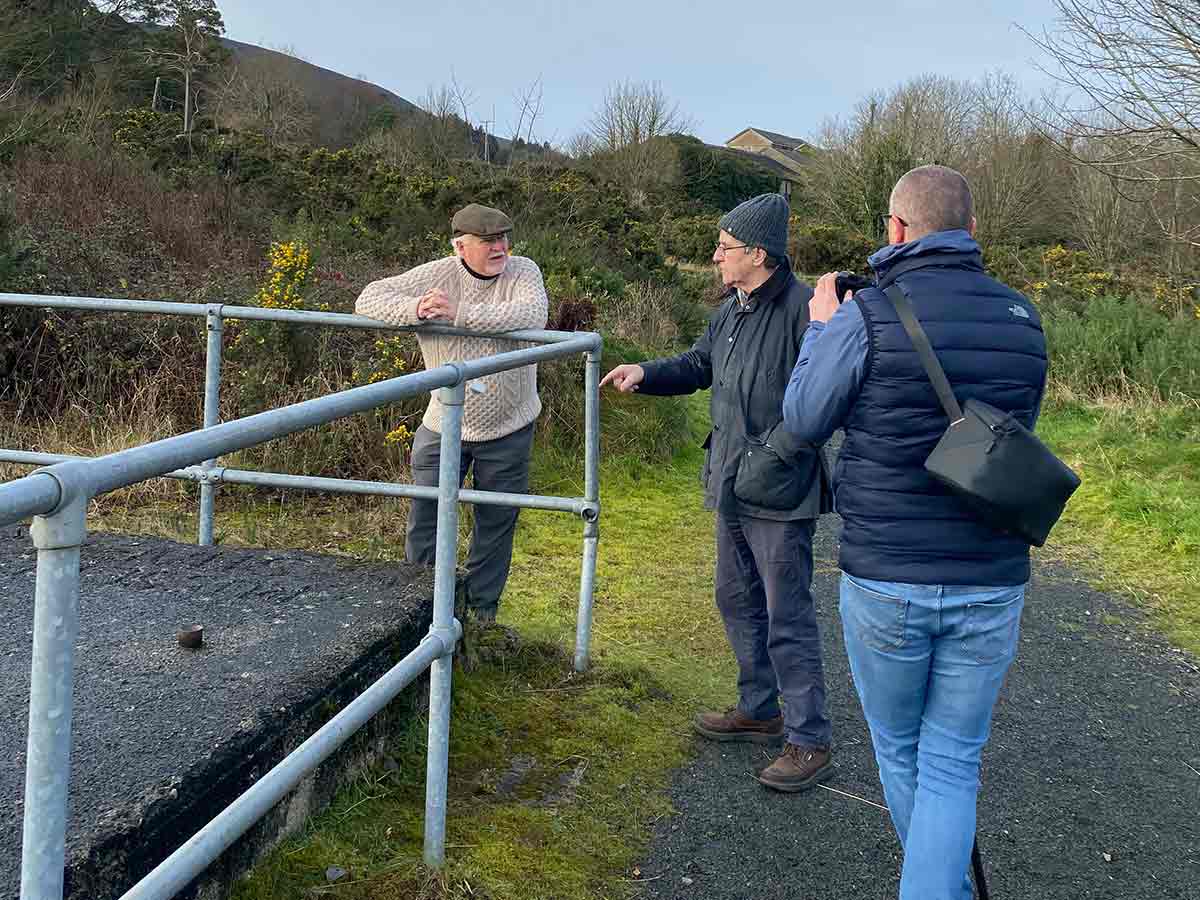 Three men outside near railing; one leans on it, another points, third takes a photo.