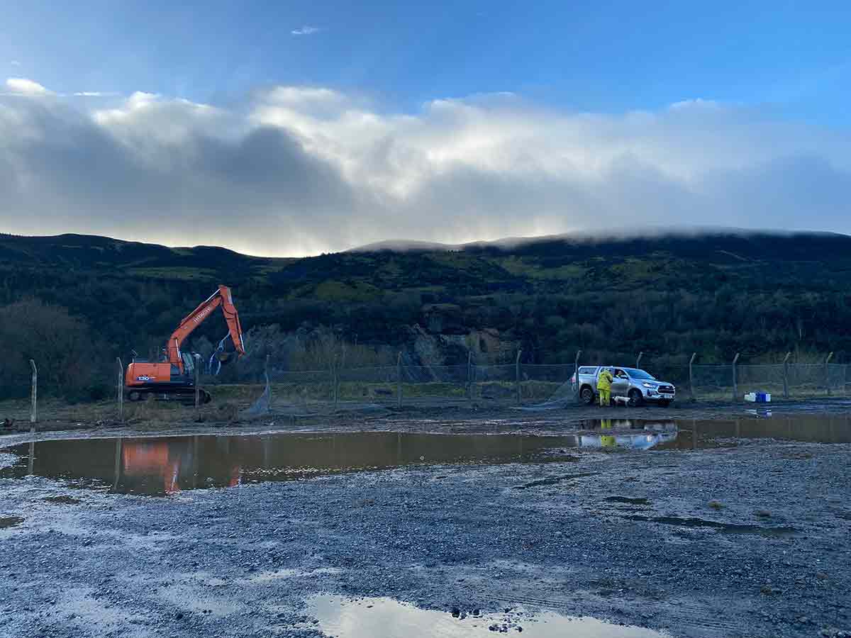 Construction site with an orange excavator, a white pickup truck, and two workers near puddles and hills.