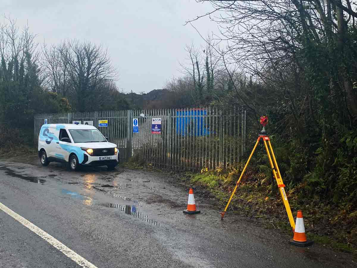Survey van parked by a fenced site entrance with surveying tripod and traffic cones on wet road.