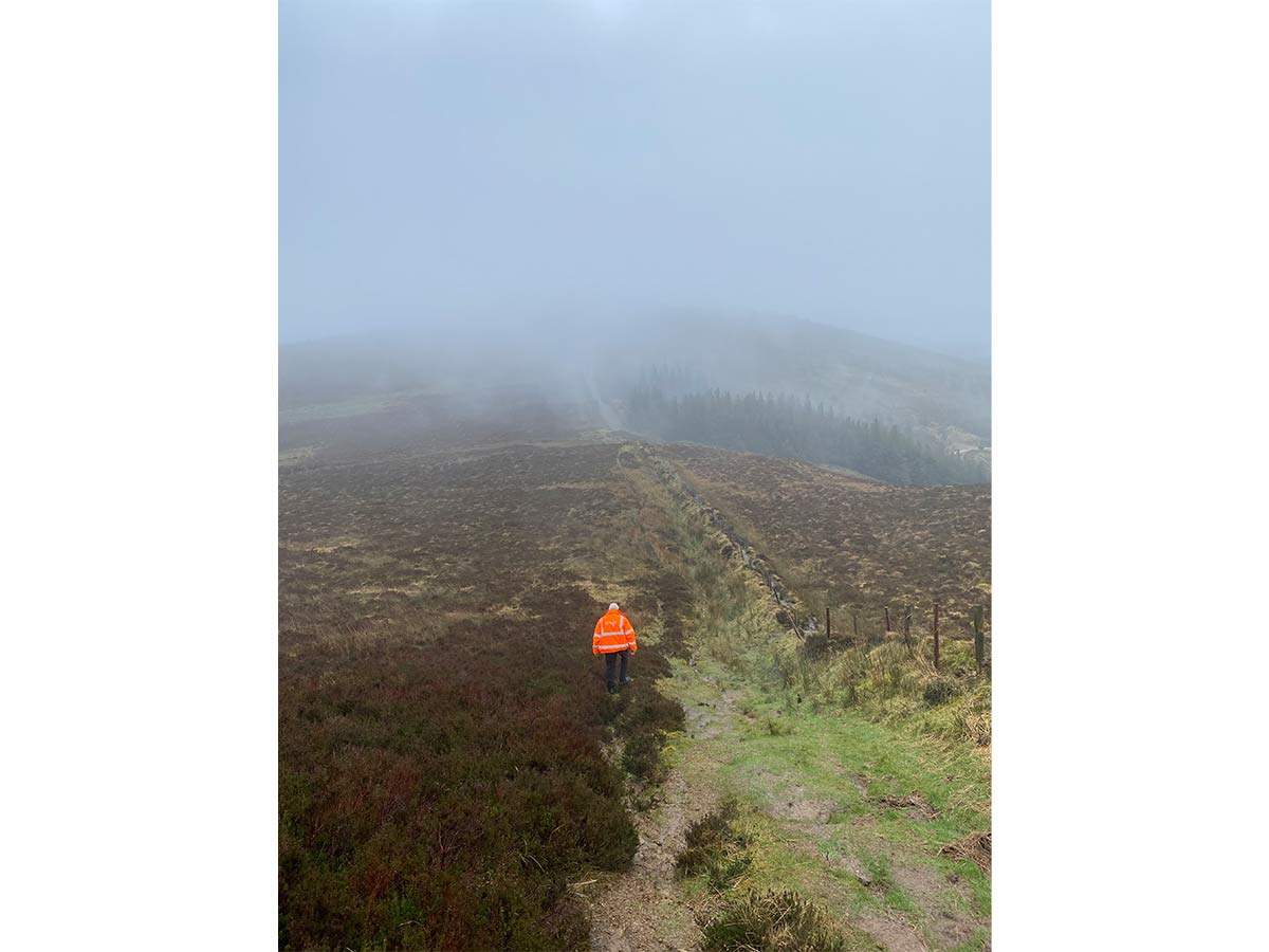 Person in bright orange jacket walking on a foggy hill path beside a stone wall and fence.