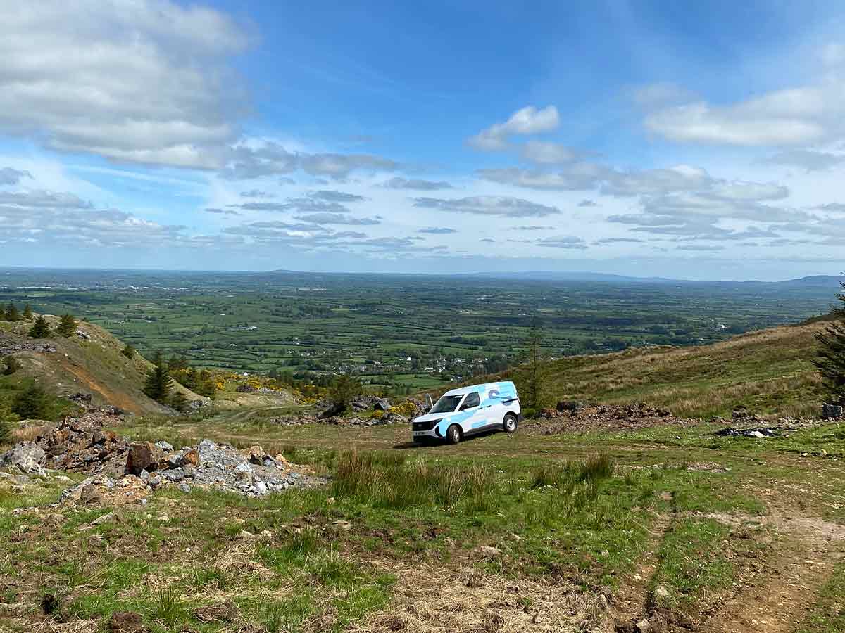 White and blue van on a dirt path in a mountainous area overlooking green fields under a blue sky.
