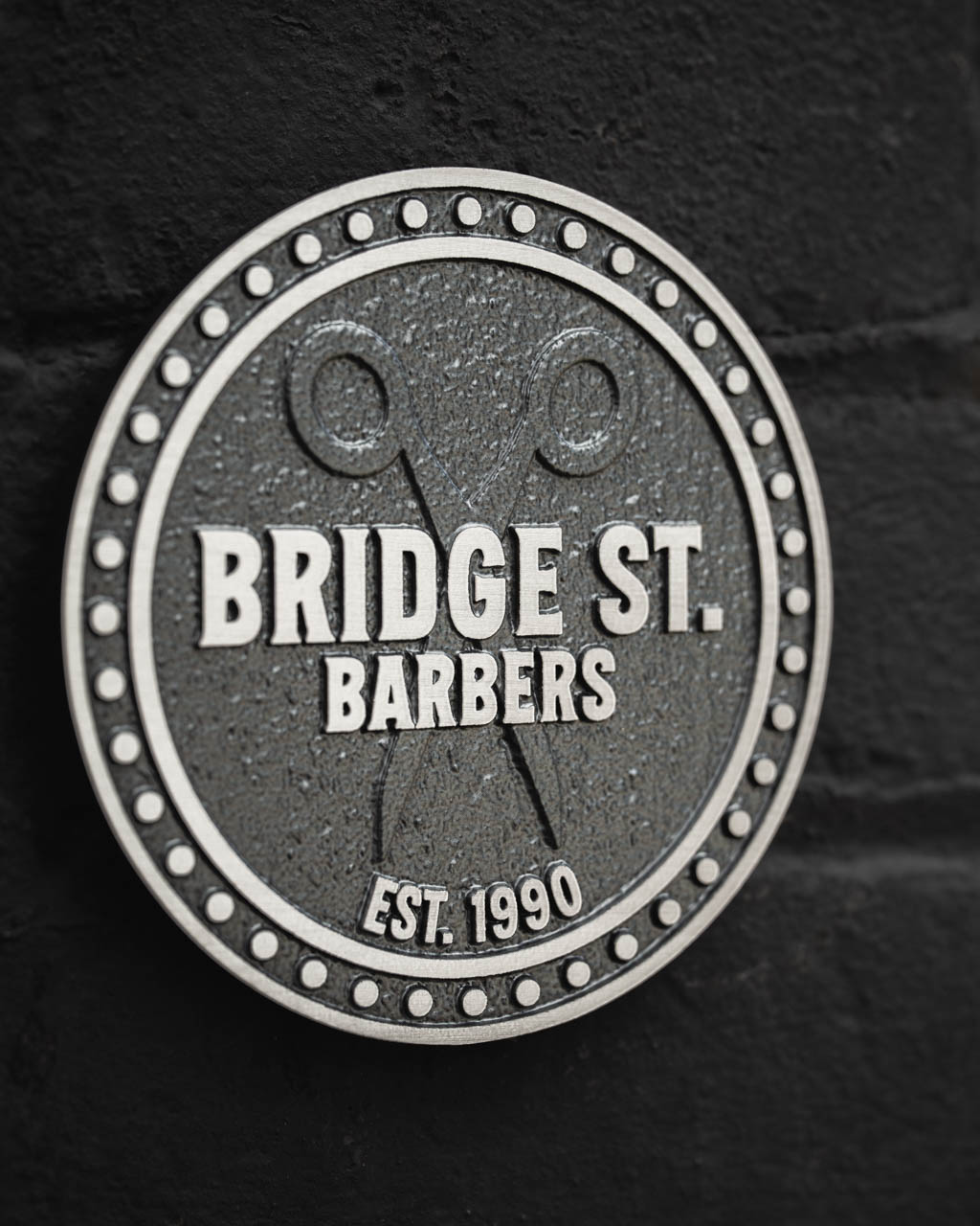 A custom circular business plaque in cast aluminium for Bridge St. Barbers, featuring raised silver lettering on a textured dark grey background. The design includes a decorative dotted border, crossed scissors motif, and the establishment date Est. 1990, offering a premium, long-lasting signage solution. Ideal for businesses seeking bespoke, handcrafted plaques to enhance their storefront with timeless branding.