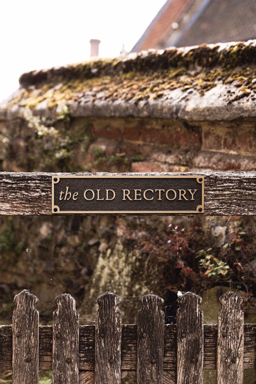 A bespoke bronze house sign mounted on a rustic stone wall, featuring the text The Old Rectory in raised gold lettering on a textured dark brown background. The plaque is rectangular with chamfered corners and elegant serif typography, blending traditional charm with refined craftsmanship. Ideal for heritage properties or personalised home signage, this custom house name sign exemplifies classic design and premium materials.