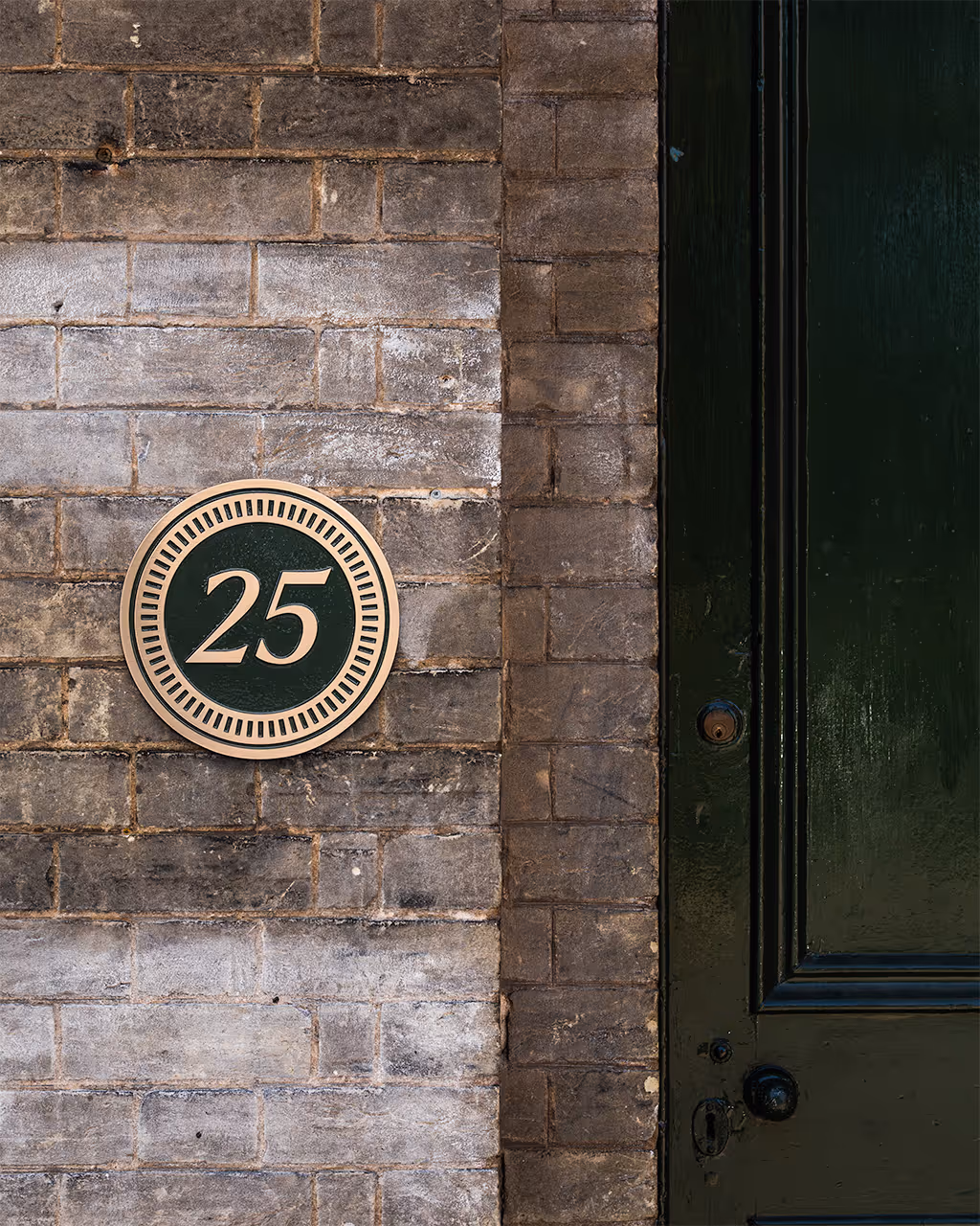 A custom luxury house sign featuring a circular bronze plaque with a painted deep green background and gold lettering. The sign displays the house number 25 in elegant raised lettering, framed in a decorative border. Crafted with precision, this bespoke house plaque combines timeless style with premium quality, making it an ideal choice for personalised home signage or custom bronze house number signs.