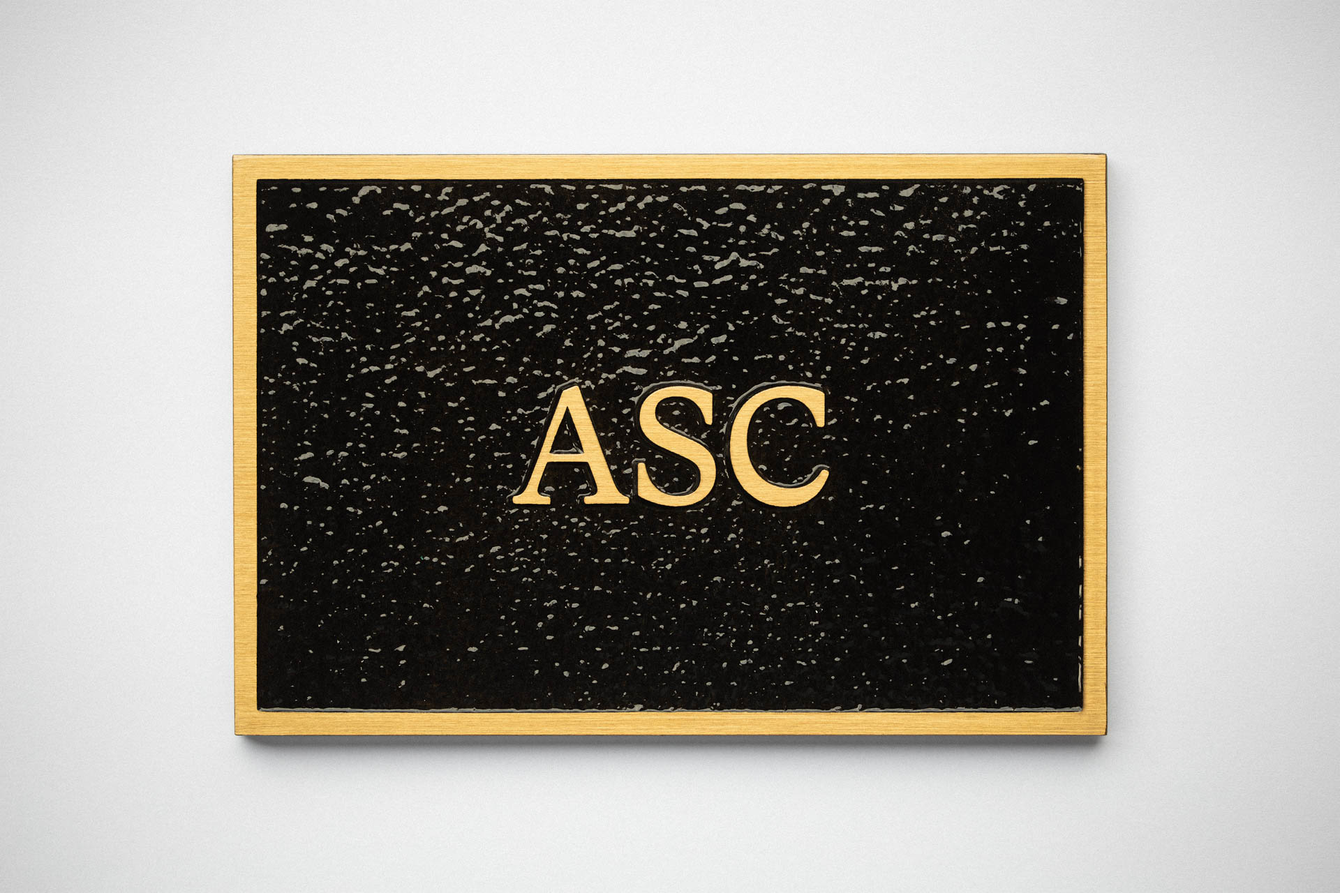 Cast bronze plaque with black mottled textured background and raised gold letters reading ASC, mounted on a light wall.