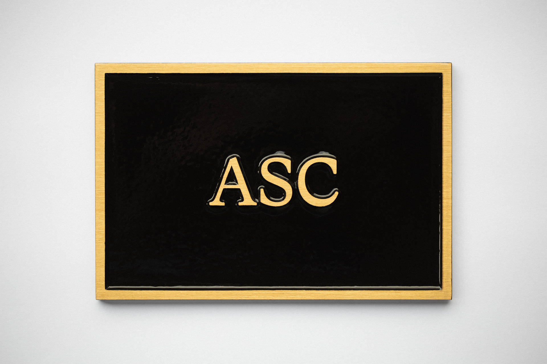 Cast bronze plaque with a smooth black background and raised gold letters reading ASC, mounted on a light wall.