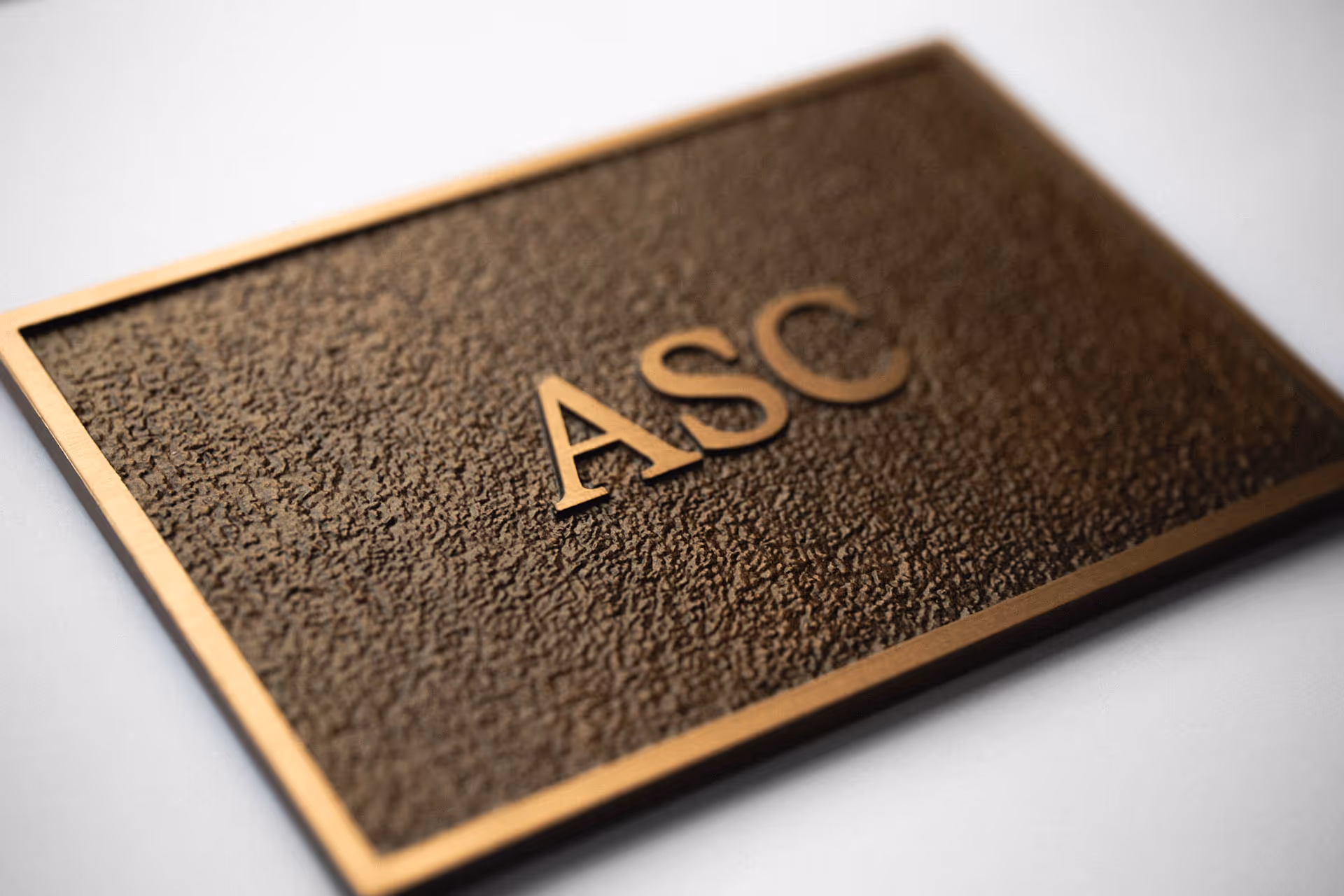 Cast bronze plaque with patinated mottled textured background and raised gold letters reading ASC, mounted on a light wall.