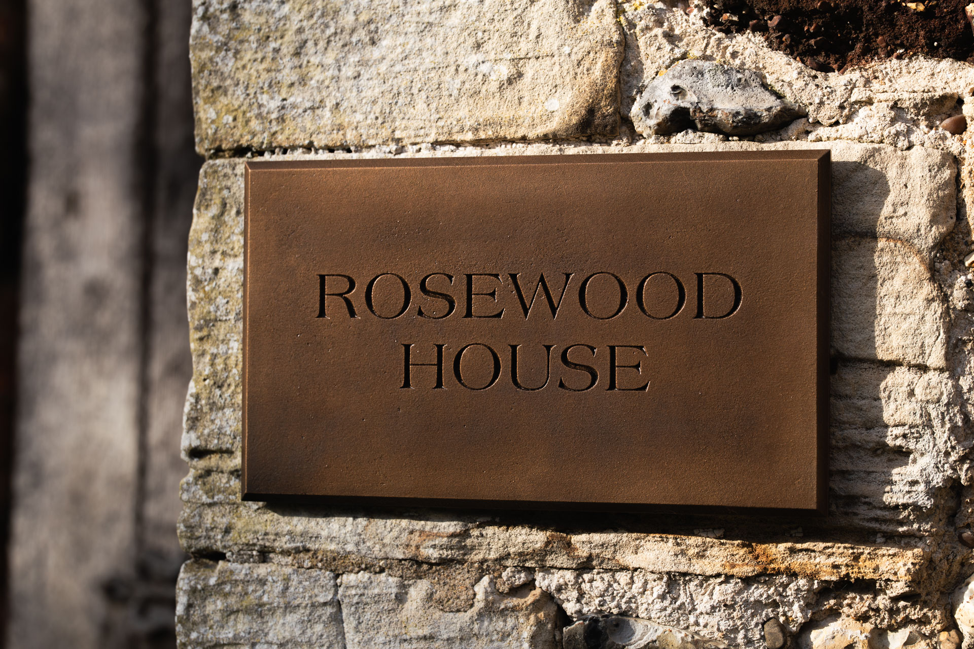 Cast bronze engraved house sign with chamfered edges and a natural patinated finish displaying “Rosewood House” mounted on a rustic stone wall.