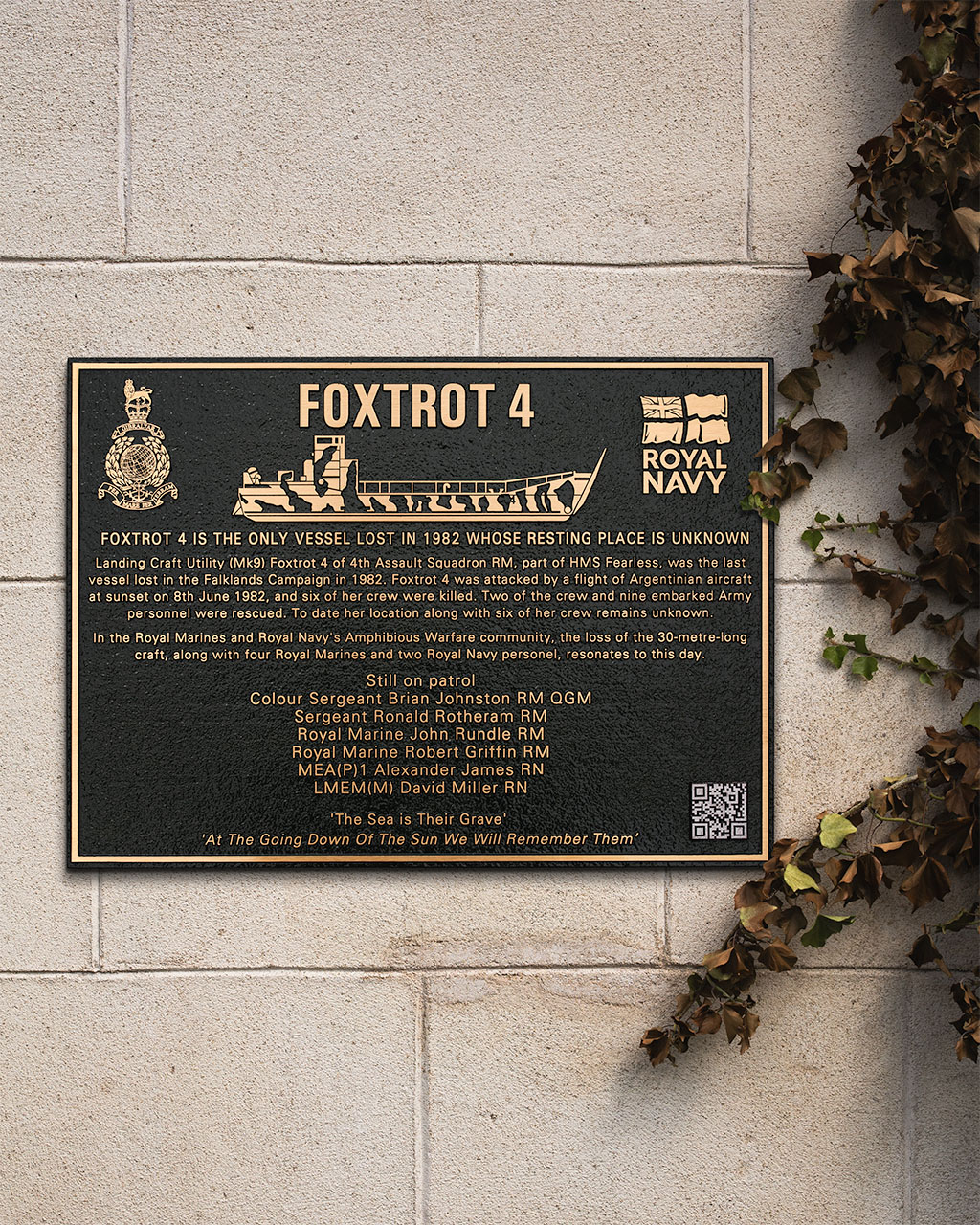 Cast bronze memorial plaque for Foxtrot 4, Royal Navy landing craft lost in the 1982 Falklands Campaign, featuring raised text, military insignias, and a relief illustration of the vessel. Mounted on a stone wall with ivy to the side, the plaque honors six fallen servicemen with a respectful tribute and engraved dedication.