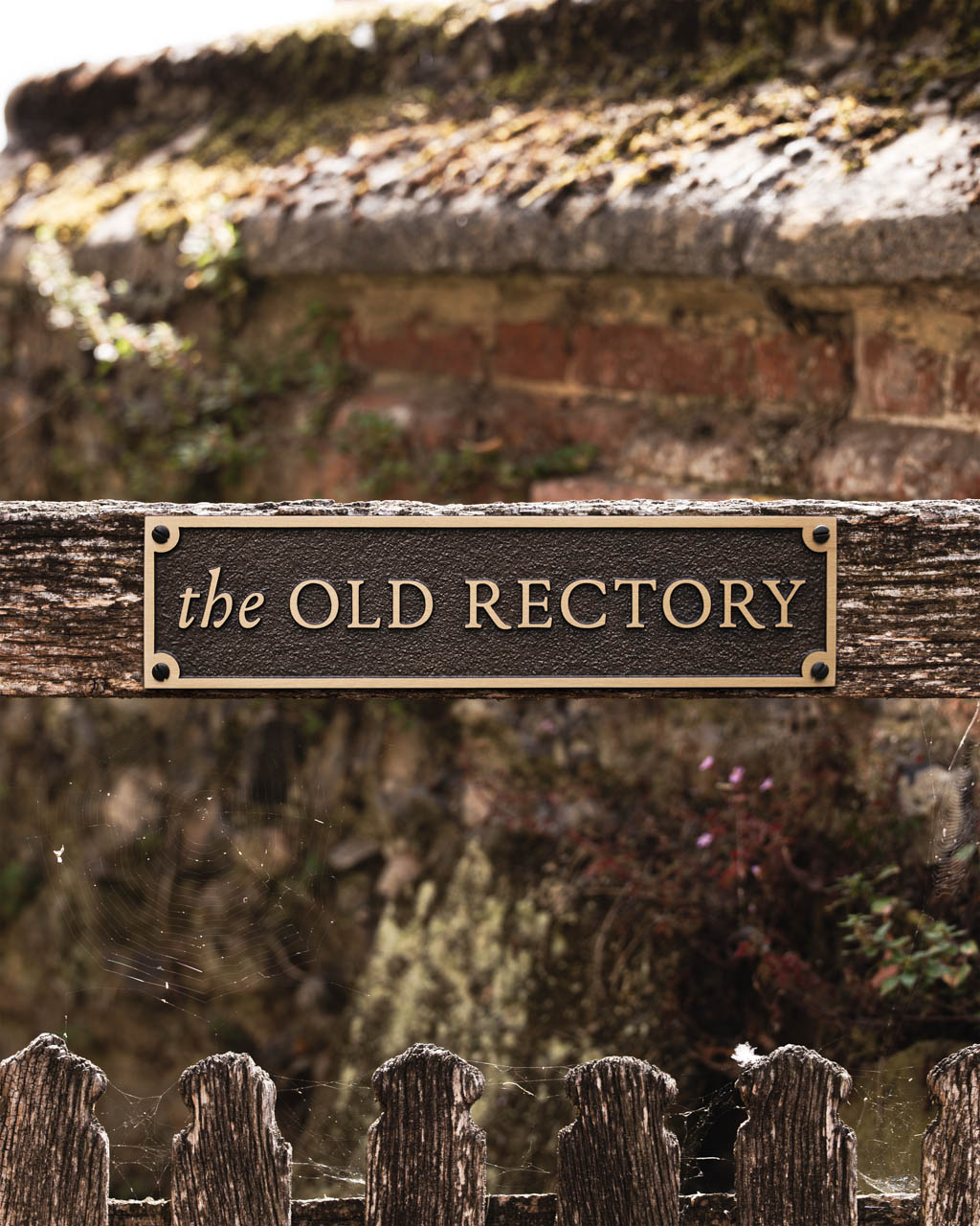 A bespoke bronze house sign mounted on a rustic gate, featuring the text The Old Rectory in raised gold lettering on a textured dark brown background. The plaque is rectangular with chamfered corners and elegant serif typography, blending traditional charm with refined craftsmanship. Ideal for heritage properties or personalised home signage, this custom house name sign exemplifies classic design and premium materials.