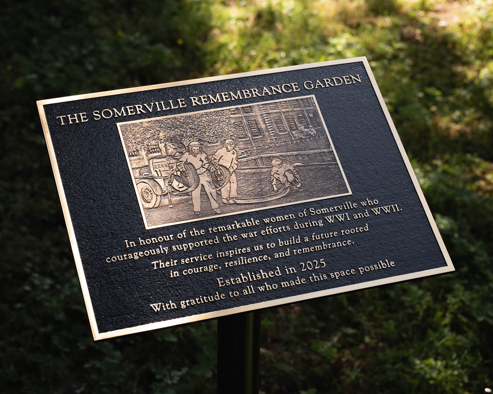 Custom cast bronze remembrance plaque in honour of the remarkable women of Somerville who courageously supported the war efforts during WWI and WWII, featuring raised gold lettering on a black background, displayed on a bespoke metal post. Handcrafted memorial signage by Anglia Sign Casting.