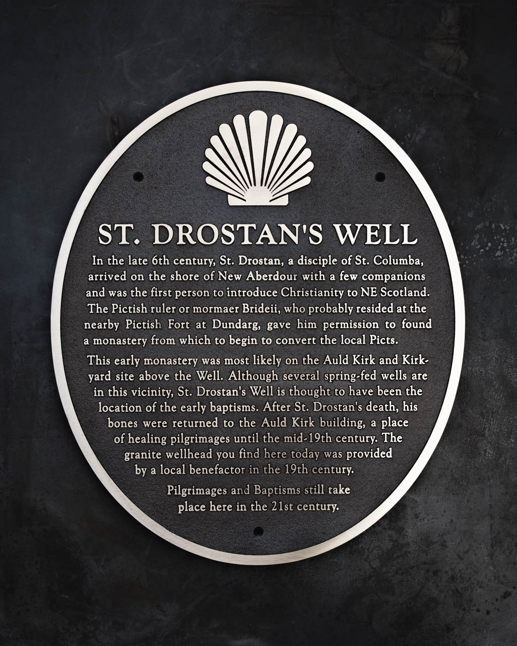 A cast bronze historical plaque titled St Drostan’s Well, handcrafted for New Aberdour. The plaque features raised lettering and a decorative scallop shell motif, symbolising pilgrimage and baptism. It recounts the story of St Drostan, a disciple of St Columba, who introduced Christianity to northeast Scotland in the late 6th century and founded a monastery near the well. The design includes a patinated background that contrasts with polished bronze highlights. Made by Anglia Sign Casting to celebrate the site’s enduring spiritual significance.