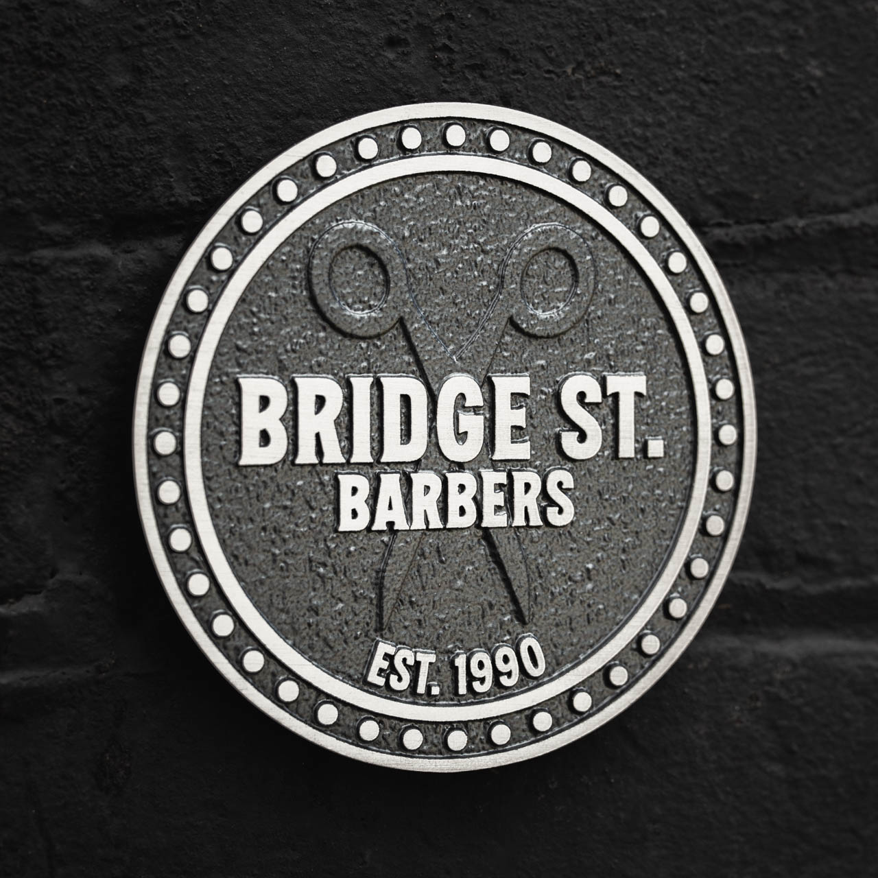 A custom circular business plaque in cast aluminium for Bridge St. Barbers, featuring raised silver lettering on a textured dark grey background. The design includes a decorative dotted border, crossed scissors motif, and the establishment date Est. 1990, offering a premium, long-lasting signage solution. Ideal for businesses seeking bespoke, handcrafted plaques to enhance their storefront with timeless branding.