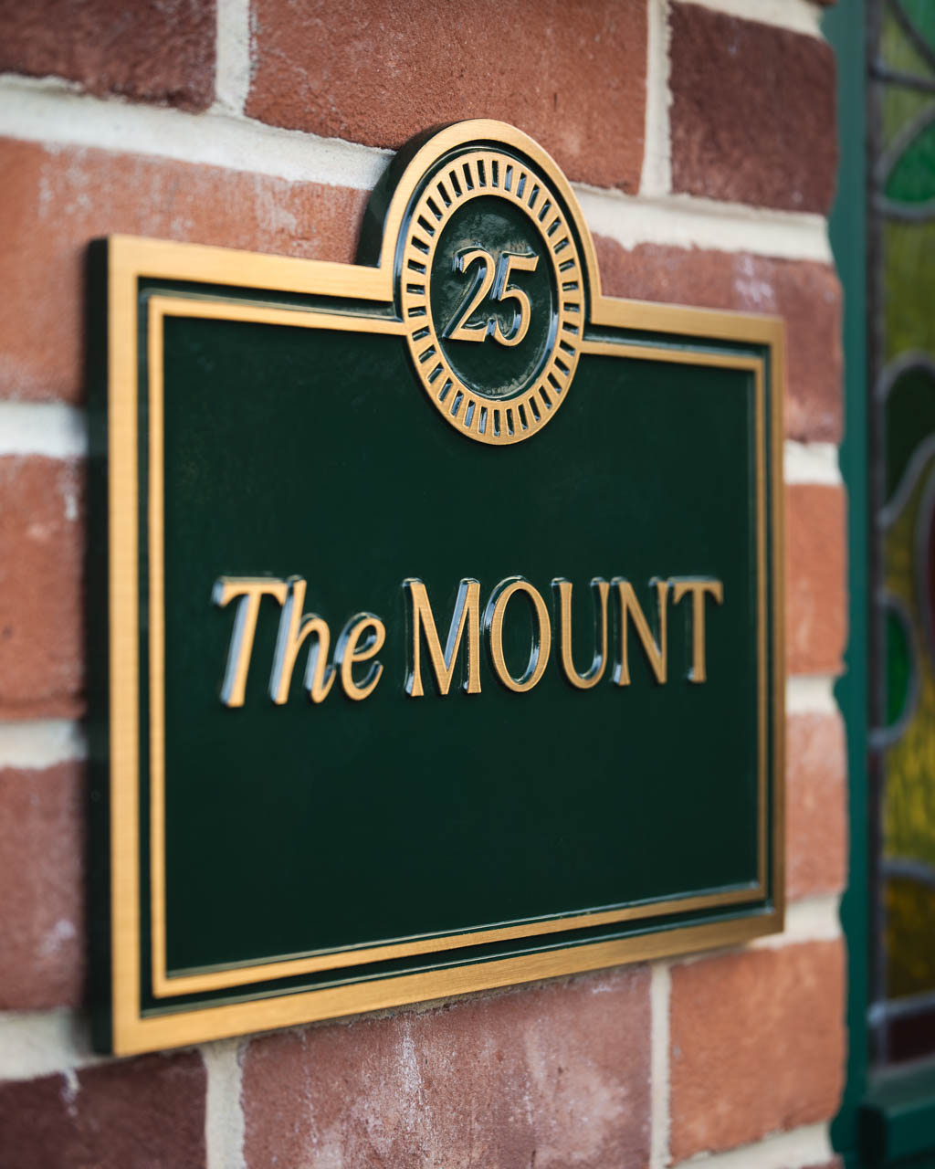 A custom luxury house sign featuring a rectangular bronze plaque with a painted deep green background and gold lettering. The sign displays the house name The Mount in elegant raised lettering, with the number 25 framed in a circular motif at the top. Crafted with precision, this bespoke house plaque combines timeless style with premium quality, making it an ideal choice for personalised home signage or custom bronze house number signs.
