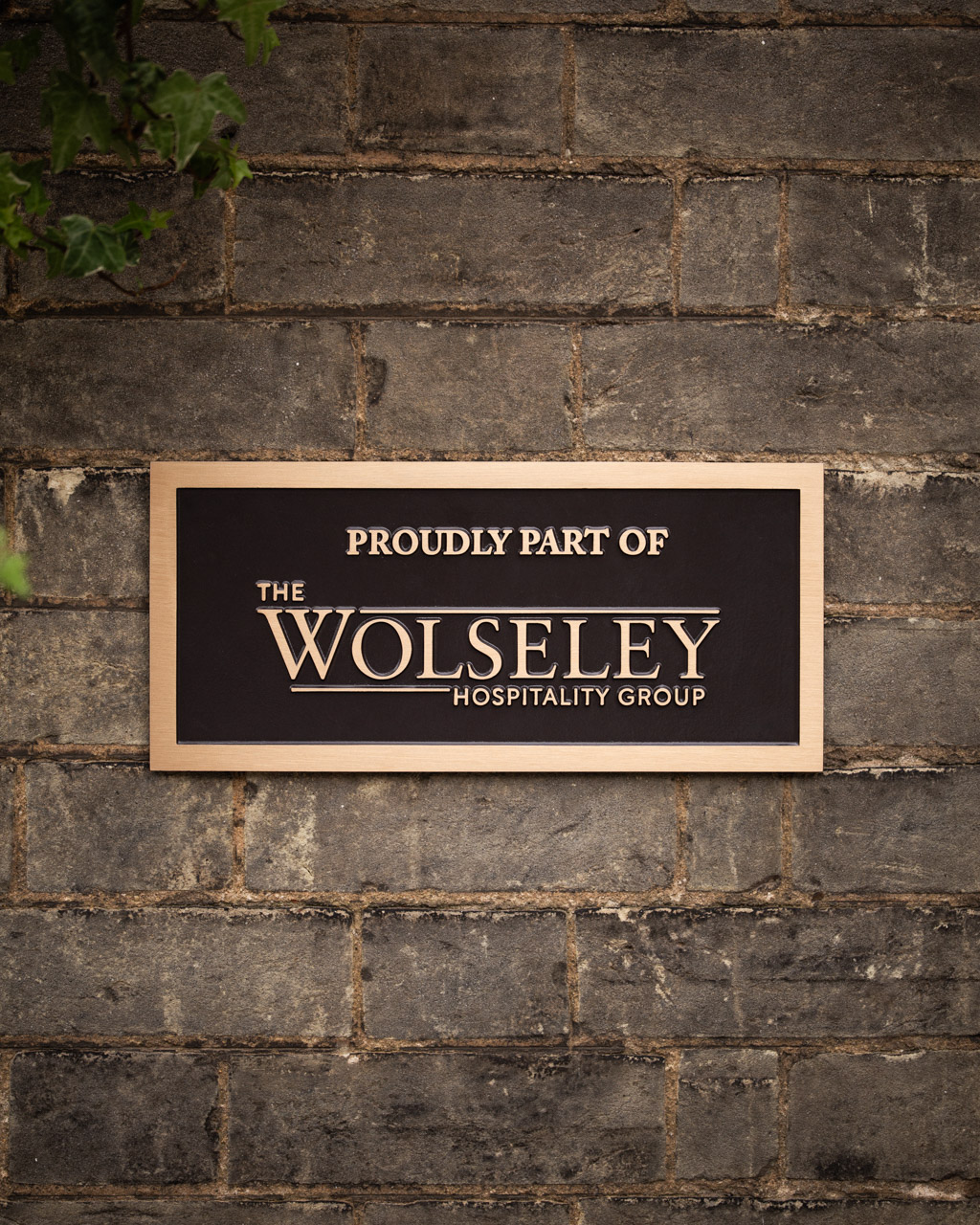 A luxury cast bronze business plaque with raised lettering reading Proudly part of The Wolseley Hospitality Group. The plaque features a brushed bronze border and polished text on a painted brown background, mounted on a stone wall. Ideal for premium hospitality brands and businesses seeking high-quality, bespoke signage to enhance their brand presence.