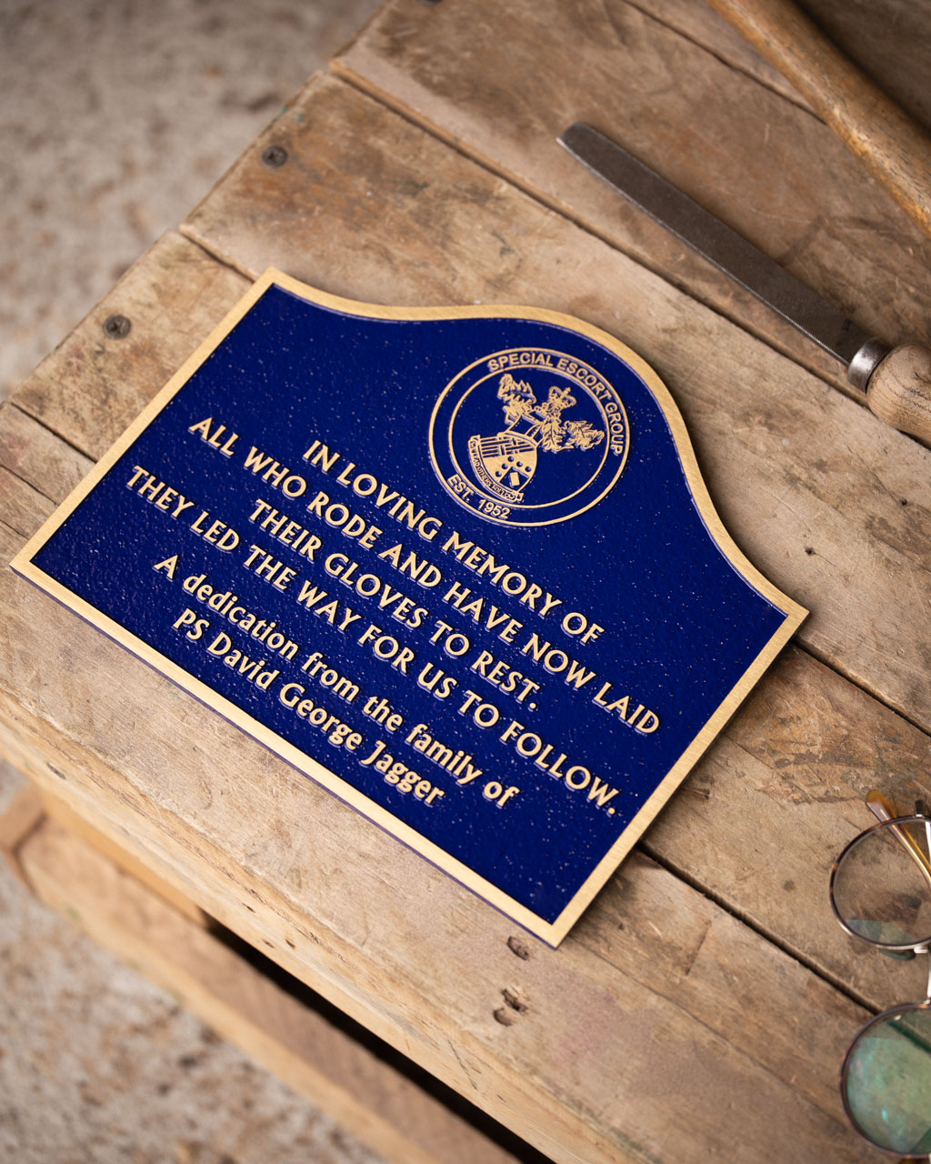 A curved bronze remembrance plaque with a rich blue background, commemorating those who rode for the Special Escort Group. The plaque features gold lettering and the group's emblem at the top, with a dedication from the family of PS David George Jagger. Crafted by Anglia Sign Casting, the plaque honours the memory of riders who have laid their gloves to rest.