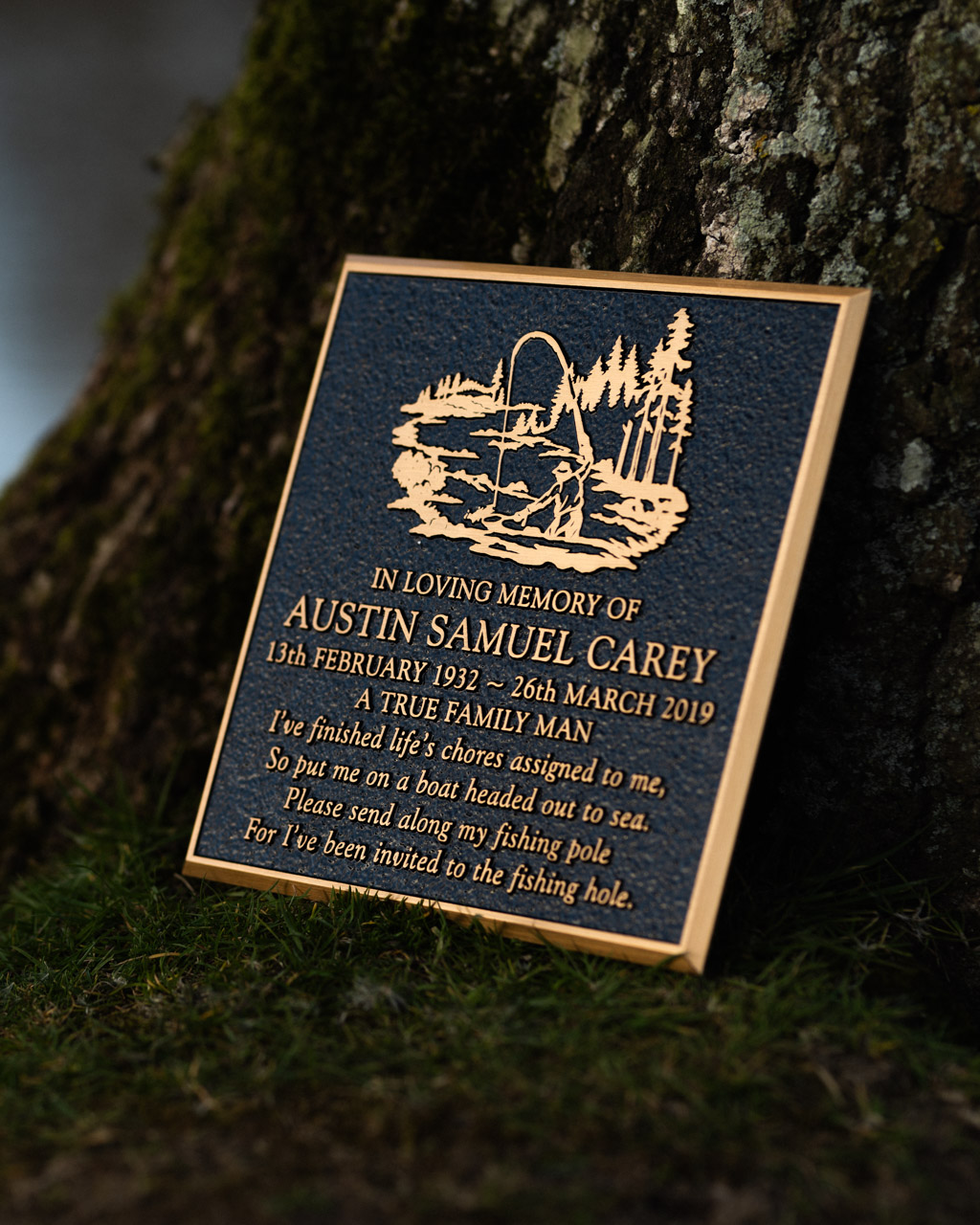 A cast bronze memorial plaque honouring Austin Samuel Carey, featuring raised lettering and a custom motif of a fisherman casting a line by a lakeside with trees in the background. The plaque's textured background contrasts with the polished lettering and artwork, creating a striking tribute to a loved one with a passion for fishing. Made by Anglia Sign Casting.