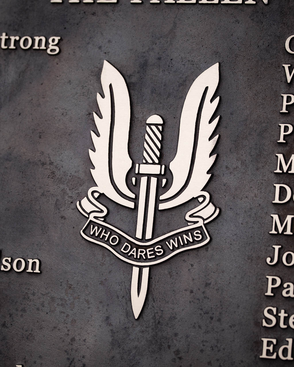 Close-up of a cast bronze military emblem featuring the iconic Special Air Service (SAS) badge with a winged dagger and the motto "Who Dares Wins." Raised bronze lettering and logo contrast against a dark patinated background, commemorating fallen servicemen with a timeless, dignified tribute.