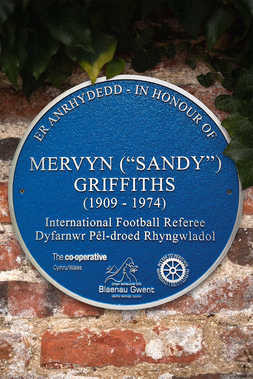 Bespoke blue heritage plaque honouring Mervyn ('Sandy') Griffiths (1909–1974), an international football referee. Featuring raised white lettering in both Welsh and English, this cast aluminium plaque is mounted on a brick wall and commissioned by Blaenau Gwent County Borough Council. Designed for historical recognition, it highlights notable figures and their contributions to Welsh heritage.