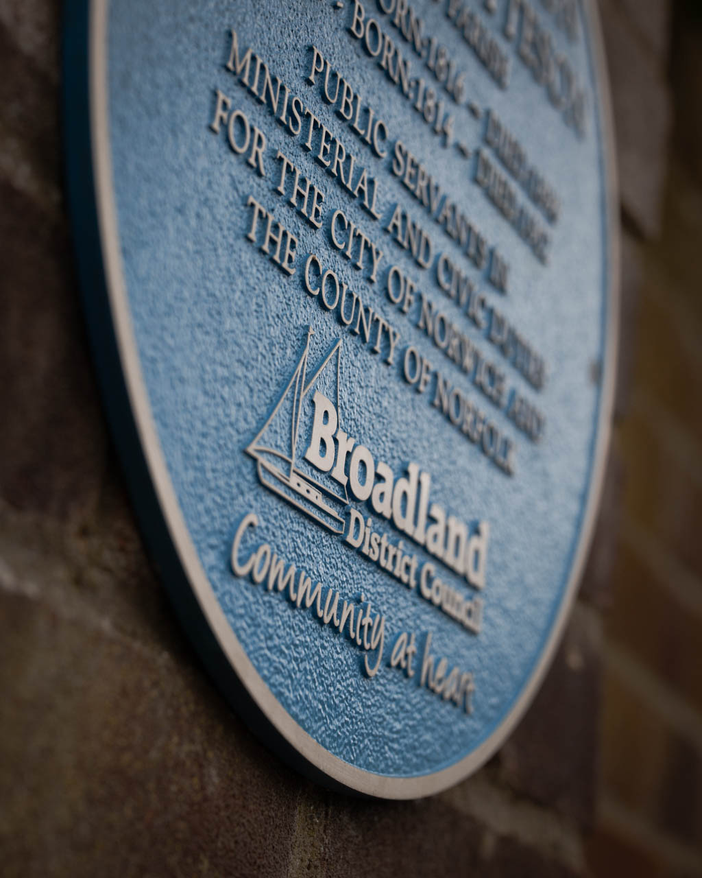 Circular blue heritage plaque commemorating Henry Patteson and John Patteson, public servants in ministerial and civic duties for Norwich and Norfolk. Affixed to a brick wall, this cast aluminium plaque features raised silver lettering and is commissioned by Broadland District Council as part of a historical recognition initiative. Designed for durability, ideal for marking historical sites and notable figures.