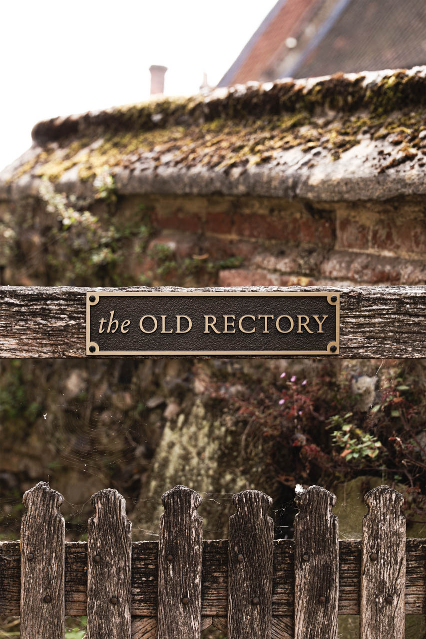 A bespoke bronze house sign mounted on a rustic stone wall, featuring the text The Old Rectory in raised gold lettering on a textured dark brown background. The plaque is rectangular with chamfered corners and elegant serif typography, blending traditional charm with refined craftsmanship. Ideal for heritage properties or personalised home signage, this custom house name sign exemplifies classic design and premium materials.