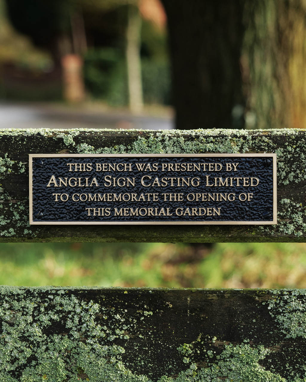 Bespoke cast bronze bench plaque with raised lettering, reading 'This bench was presented by Anglia Sign Casting Limited to commemorate the opening of this memorial garden.' Designed with a textured black background and polished gold-tone lettering, this durable and weather-resistant plaque is perfect for commemorative benches, dedication plaques, or personalised outdoor signage. Mounted on a rustic wooden bench covered in natural lichen, ideal for parks, memorial gardens, and public spaces.