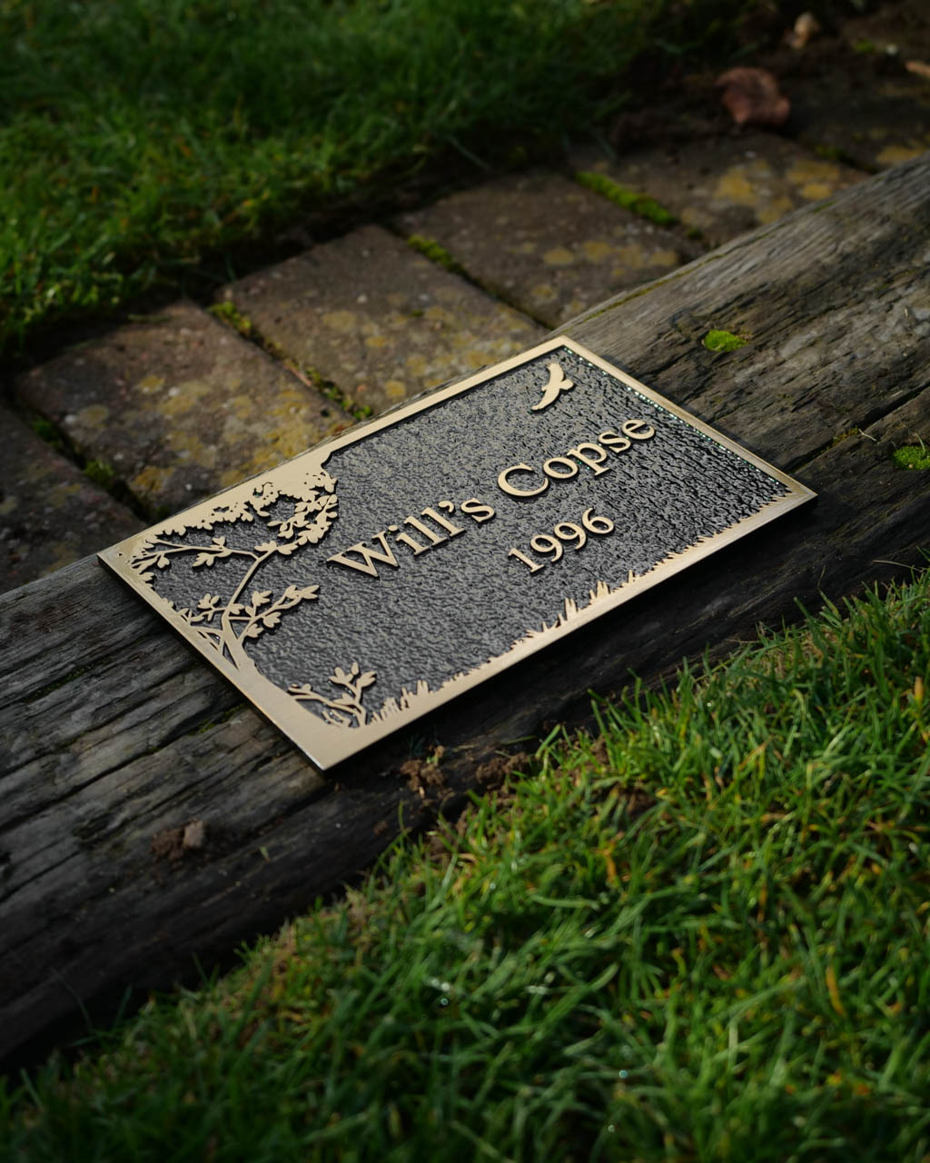 Premium cast bronze plaque with raised lettering, reading 'Will’s Copse 1996.' This bespoke outdoor nameplate features a textured background, decorative tree and bird design, and a weather-resistant finish. Ideal for garden markers, woodland dedications, or personalised outdoor signage. Displayed on a rustic wooden beam with grass and a moss-covered brick path in the background.