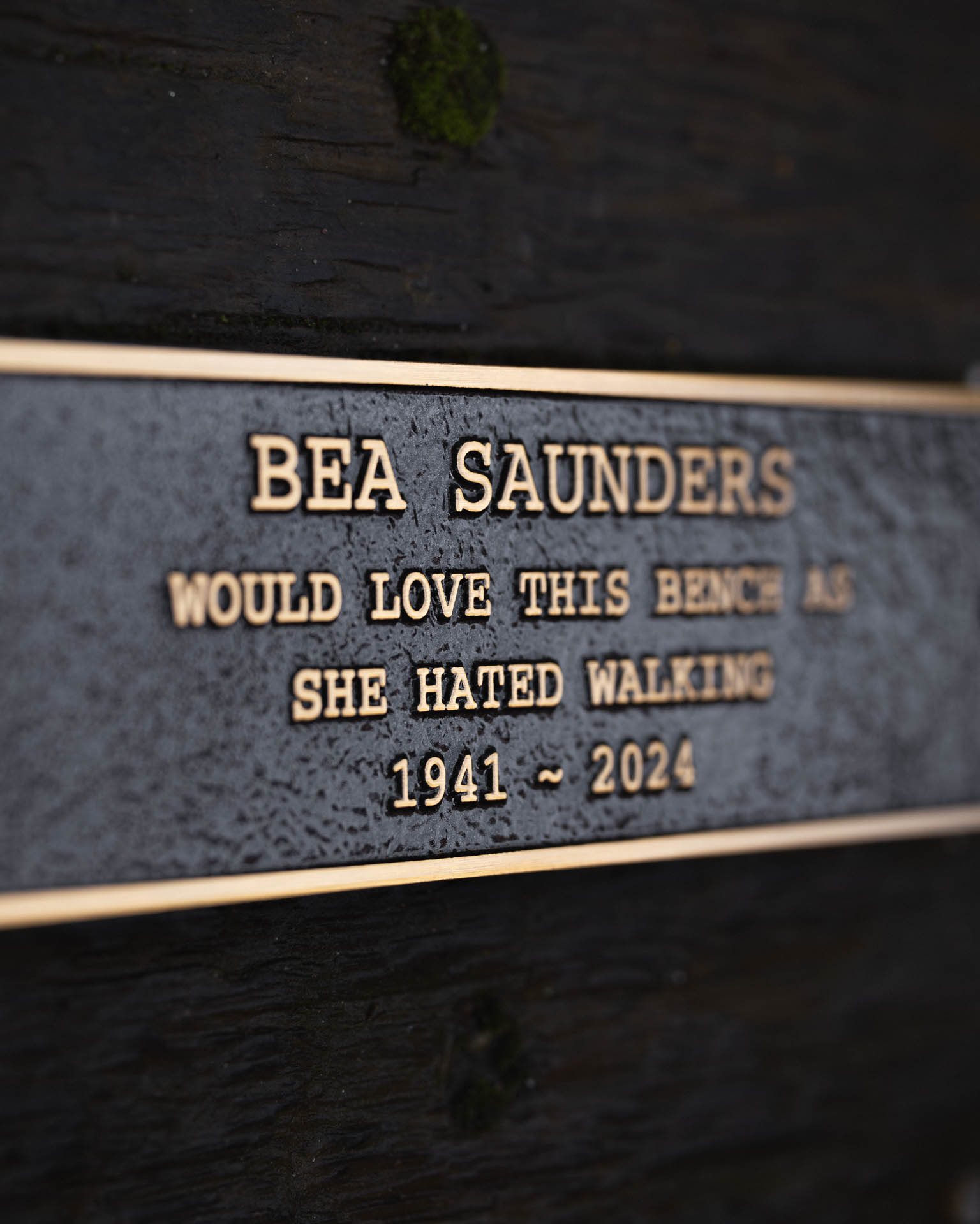 Custom cast bronze bench plaque with raised gold-tone lettering, reading 'Bea Saunders would love this bench as she hated walking 1941 - 2024.' Featuring a textured black background, durable weather-resistant finish, and pre-drilled holes for easy mounting. This personalised dedication plaque is perfect for memorial benches, tribute markers, and outdoor seating areas in parks and gardens.