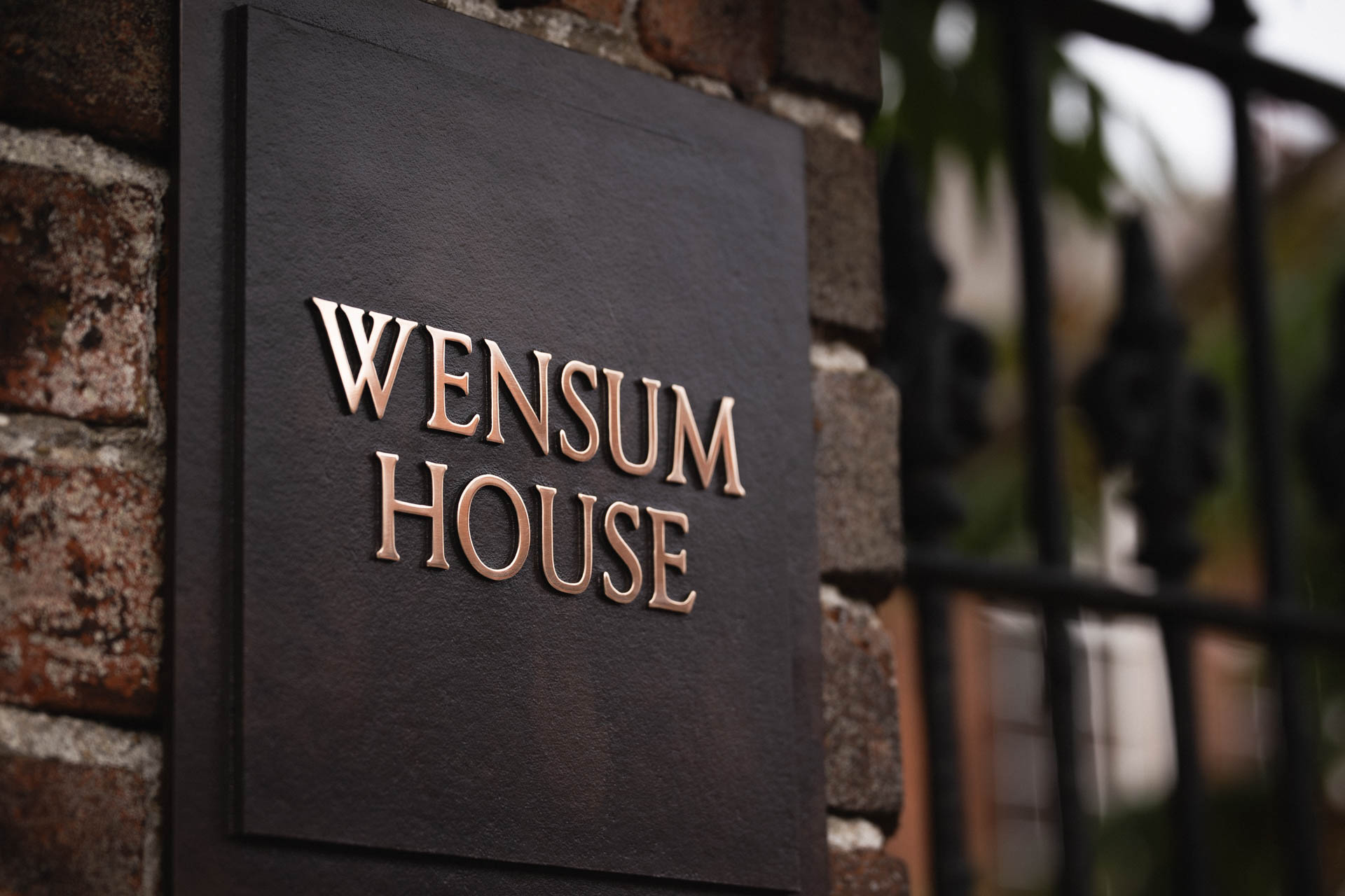 Cast bronze house sign reading “Wensum House” with raised polished bronze lettering, a dark patinated background, and a two-tiered design, mounted on a brick wall beside black wrought iron railings.