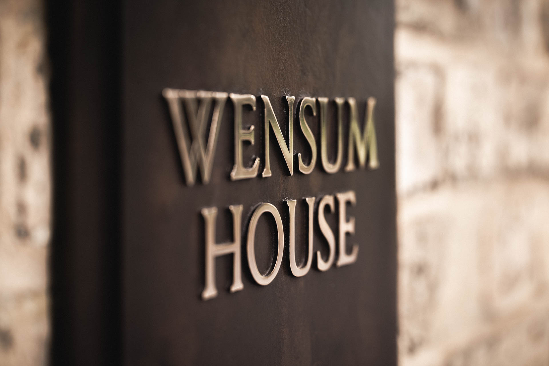 Close-up of a cast bronze house sign reading “Wensum House” with raised polished bronze lettering, dark patinated background, and two-tiered design, mounted on a light brick wall.