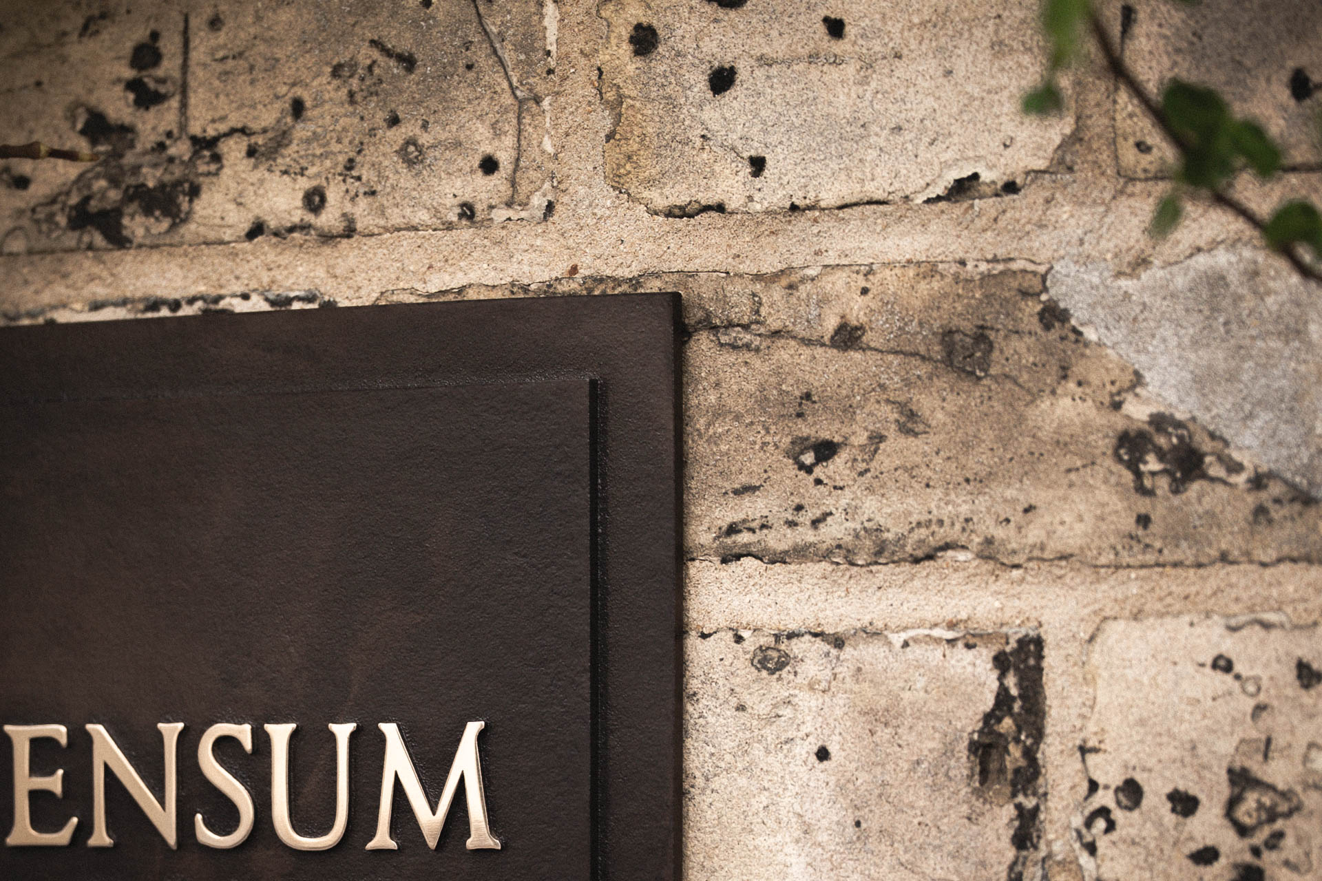 Partial view of a cast bronze house sign with raised polished bronze lettering, dark patinated background, and two-tiered design, mounted on a weathered light brick wall.